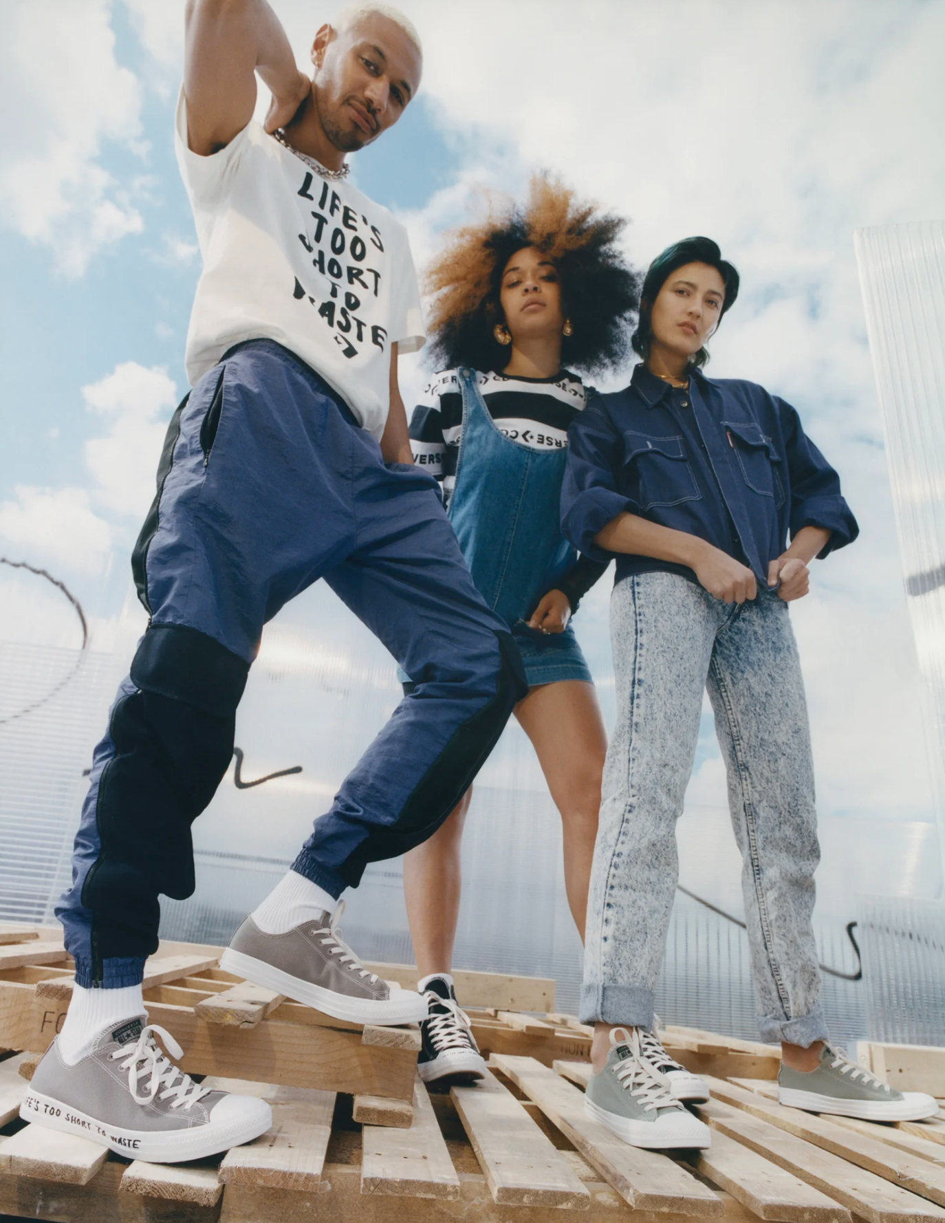 Three young adults standing on pallets outdoors against a cloudy sky, wearing casual streetwear and sneakers.