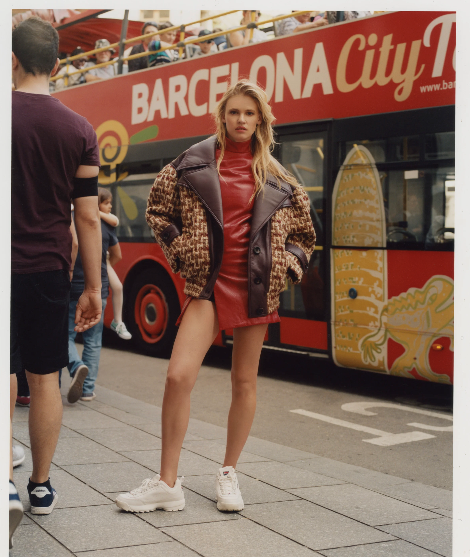 A woman with long blonde hair wearing a red dress, a patterned brown and black jacket, and white sneakers standing in front of a double-decker bus with "Barcelona City" written on it, at a busy city street.