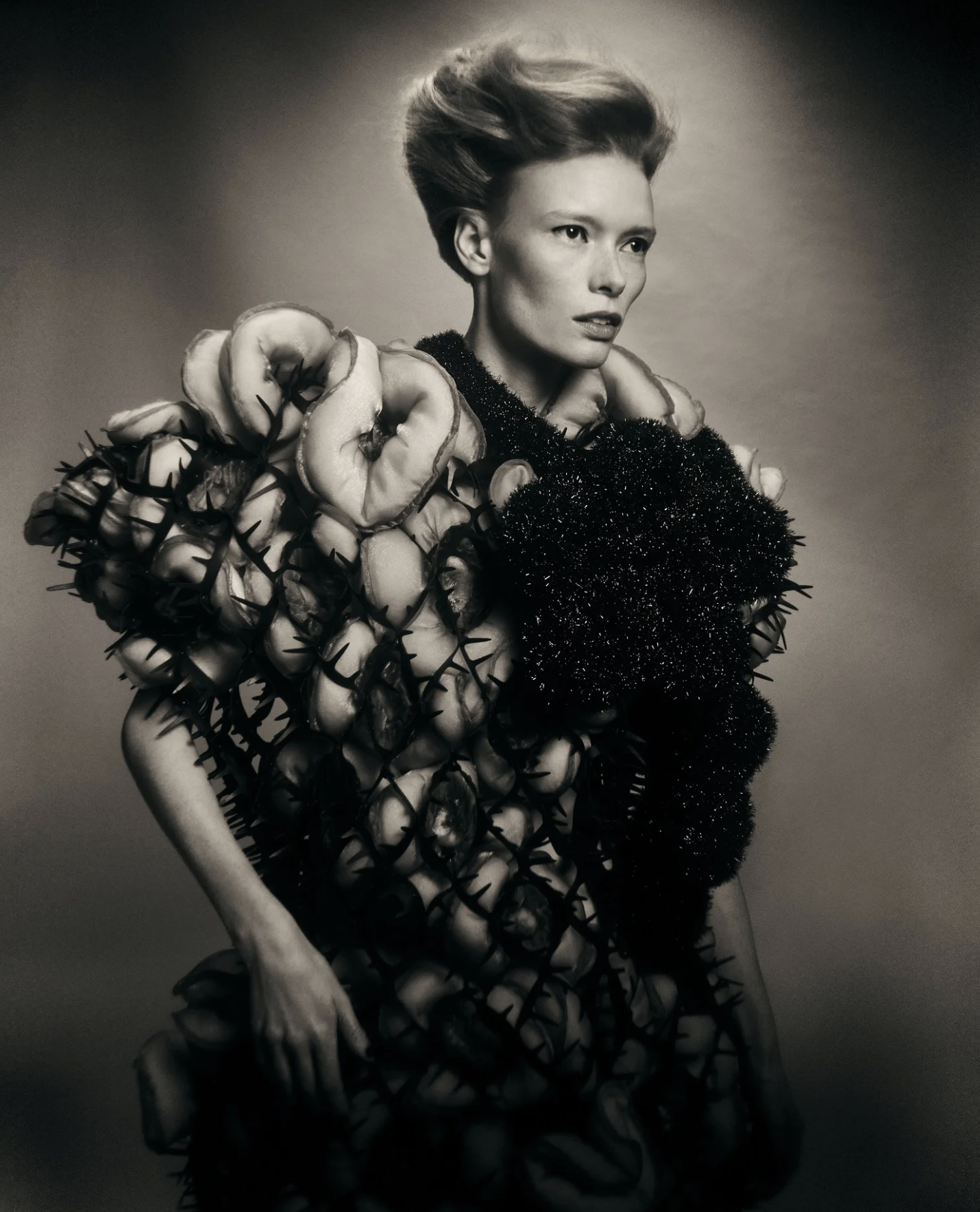 Black and white photo of a woman with voluminous hair, wearing a dress made of floral and textured fabric, standing with her hand on her hip against a plain background.