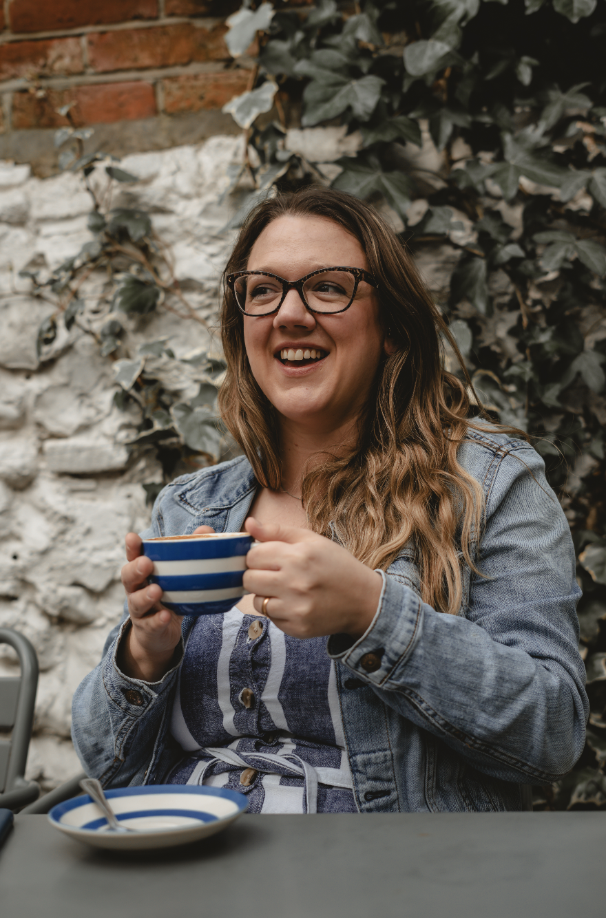 A woman with glasses, long wavy hair, and a denim jacket sitting outdoors, holding a striped blue and white mug, smiling, with ivy and a brick and stone wall in the background.