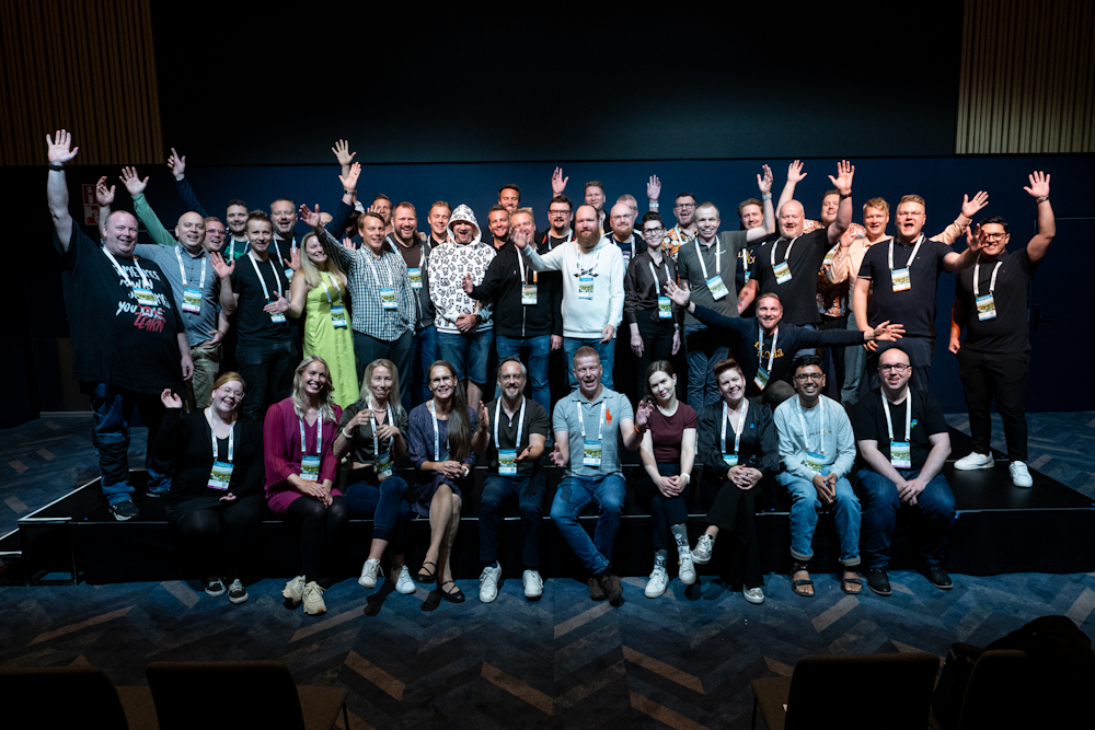 Group of people on stage at a conference, smiling and waving at the camera, wearing conference badges.