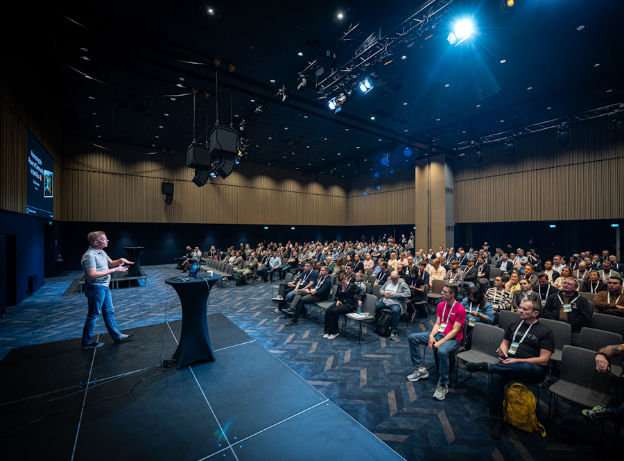 A speaker presenting on stage at a conference, with a large audience seated in front of them in a dimly lit auditorium.