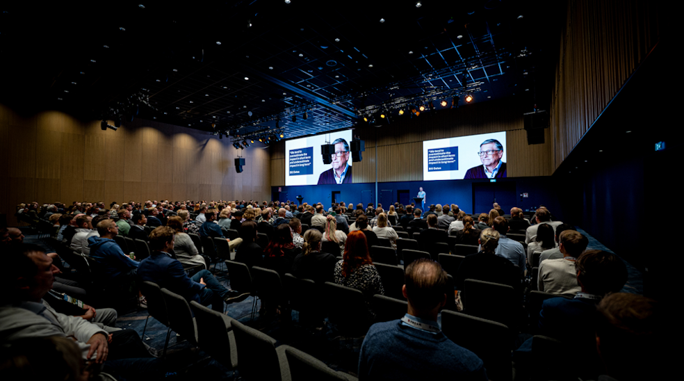 Large conference room filled with seated audience watching a speaker on stage with two large screens showing the speaker's image