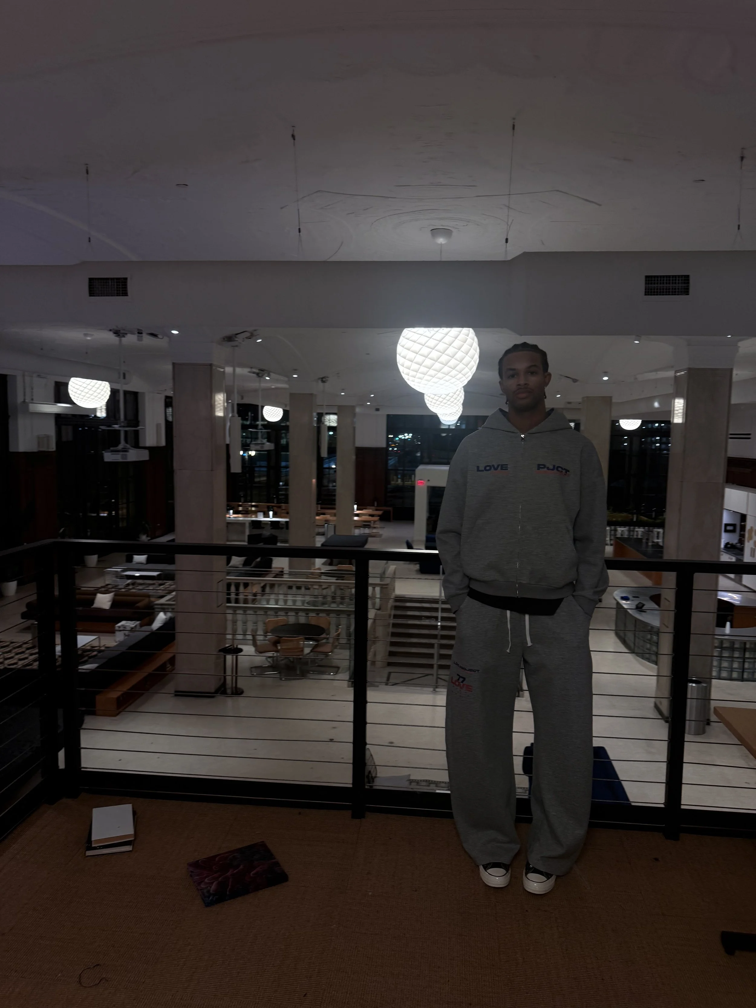 A young man wearing a gray hoodie and sweatpants standing on a balcony inside a modern, well-lit restaurant or hotel lobby at night.