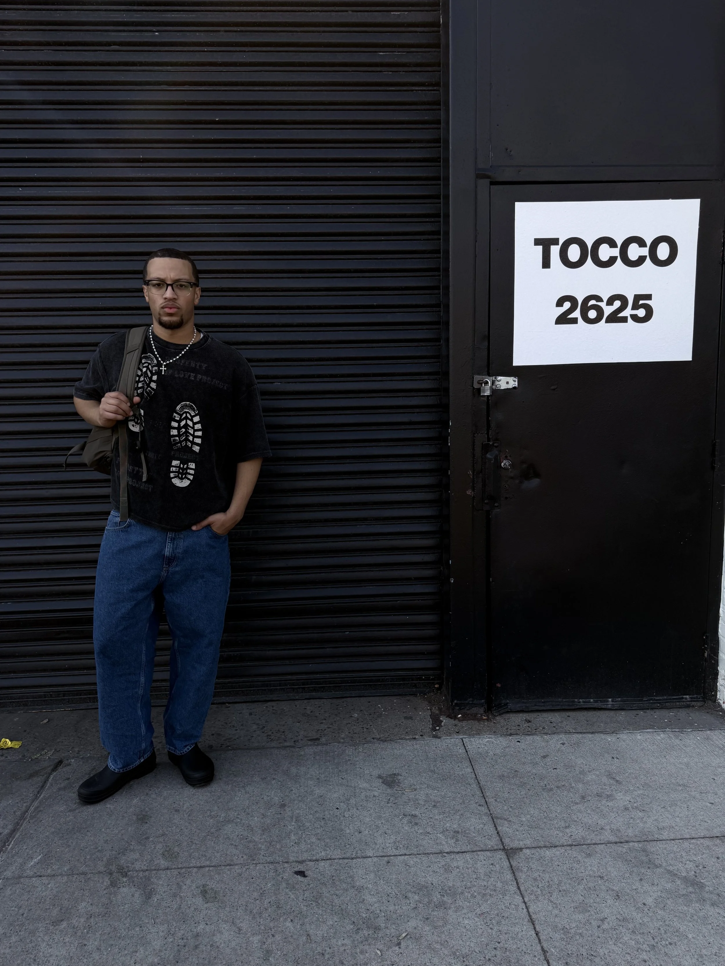A man with glasses, a goatee, and styled hair stands on a sidewalk against a black roller shutter and a black door with a sign that reads 'TOCCO 2625'. He is wearing a black t-shirt with graphic and lettering, blue jeans, and black shoes, and carries a brown bag over his shoulder.
