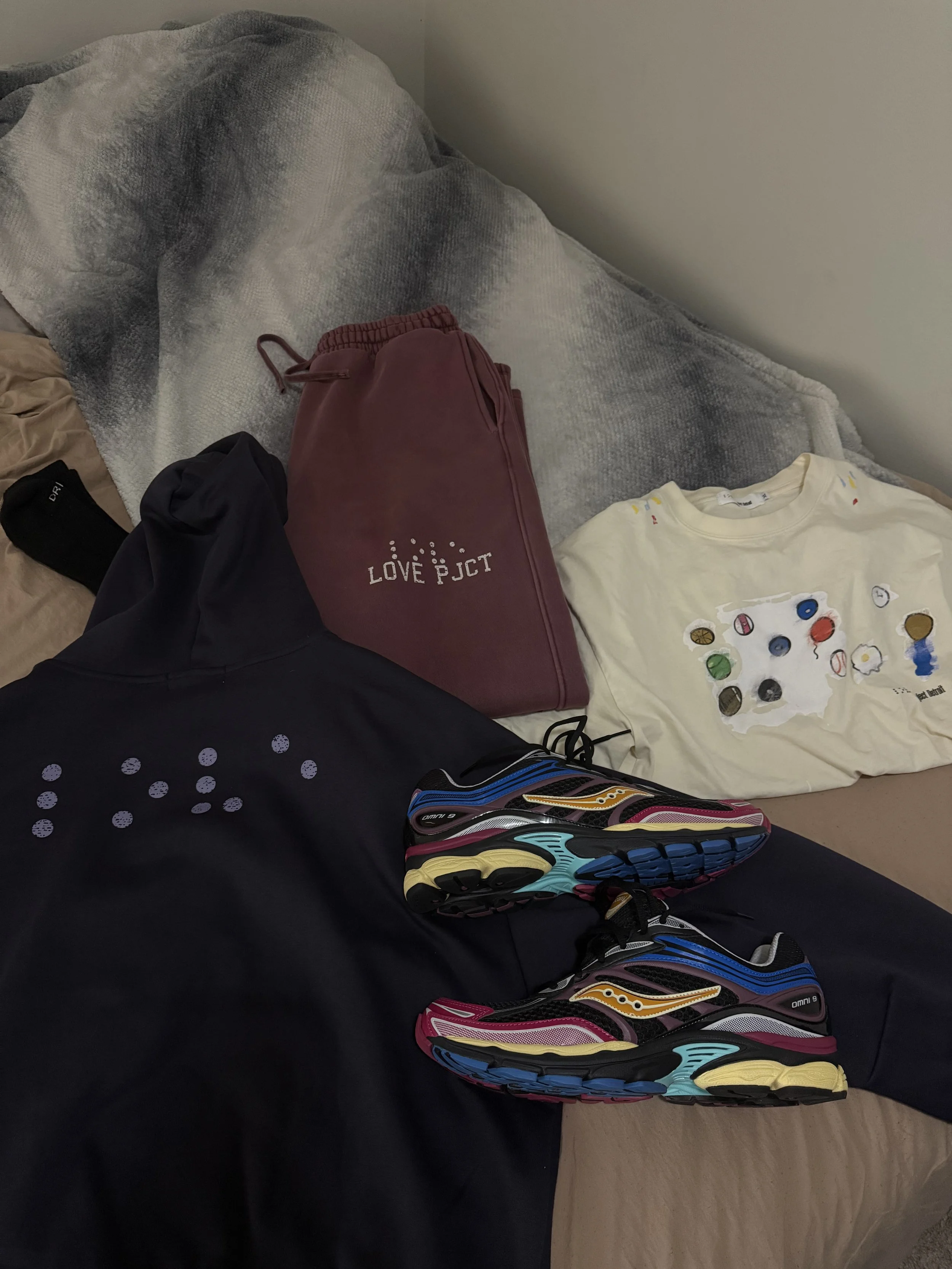 A collection of athletic clothing and sneakers laid out on a bed, including a pair of colorful running shoes, a black jacket, maroon sweatpants with 'LOVE PICT' printed on them, a black wristband, a beige t-shirt with paint splotches, and a gray blanket.