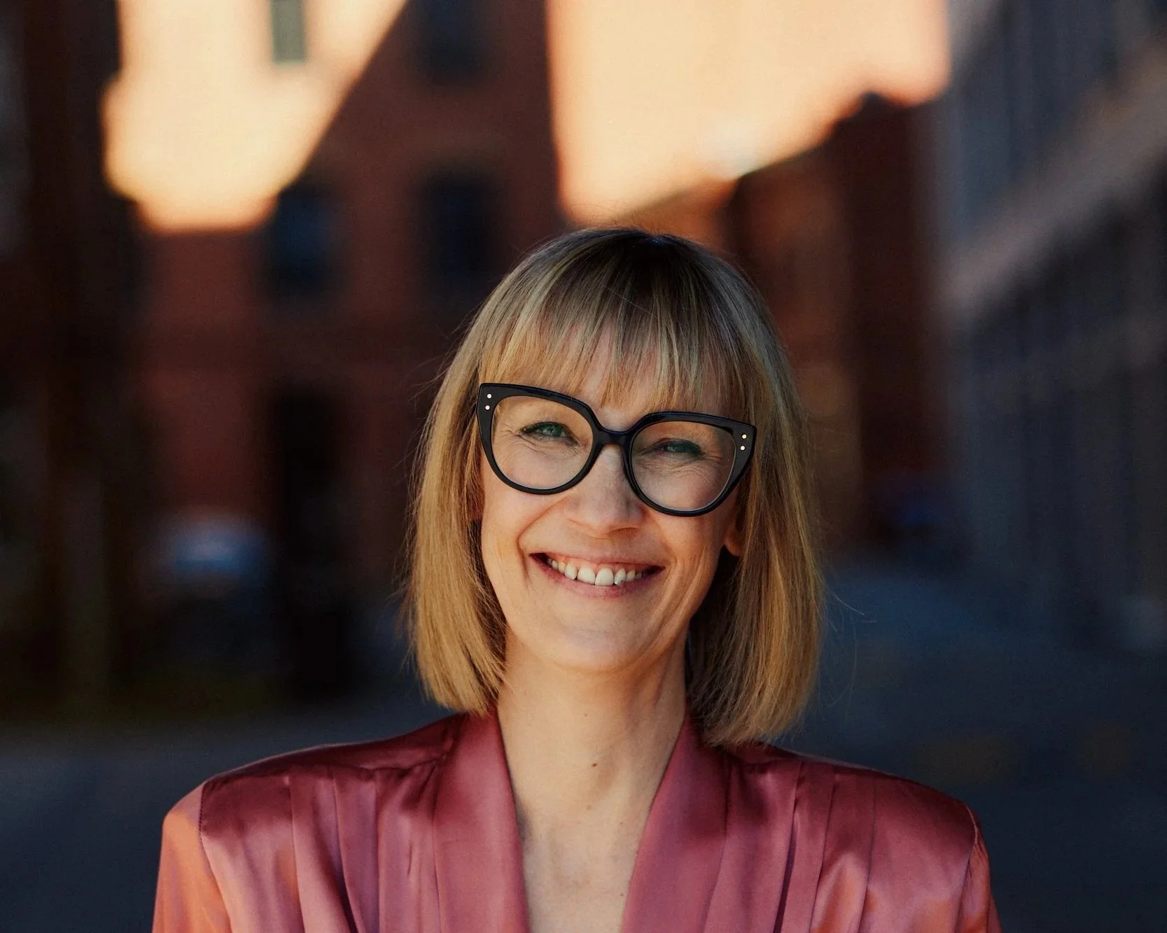 Fröhliche Frau mit braunen Haaren, Brille und rotem Kleid vor urbanem Hintergrund bei Sonnenuntergang.
