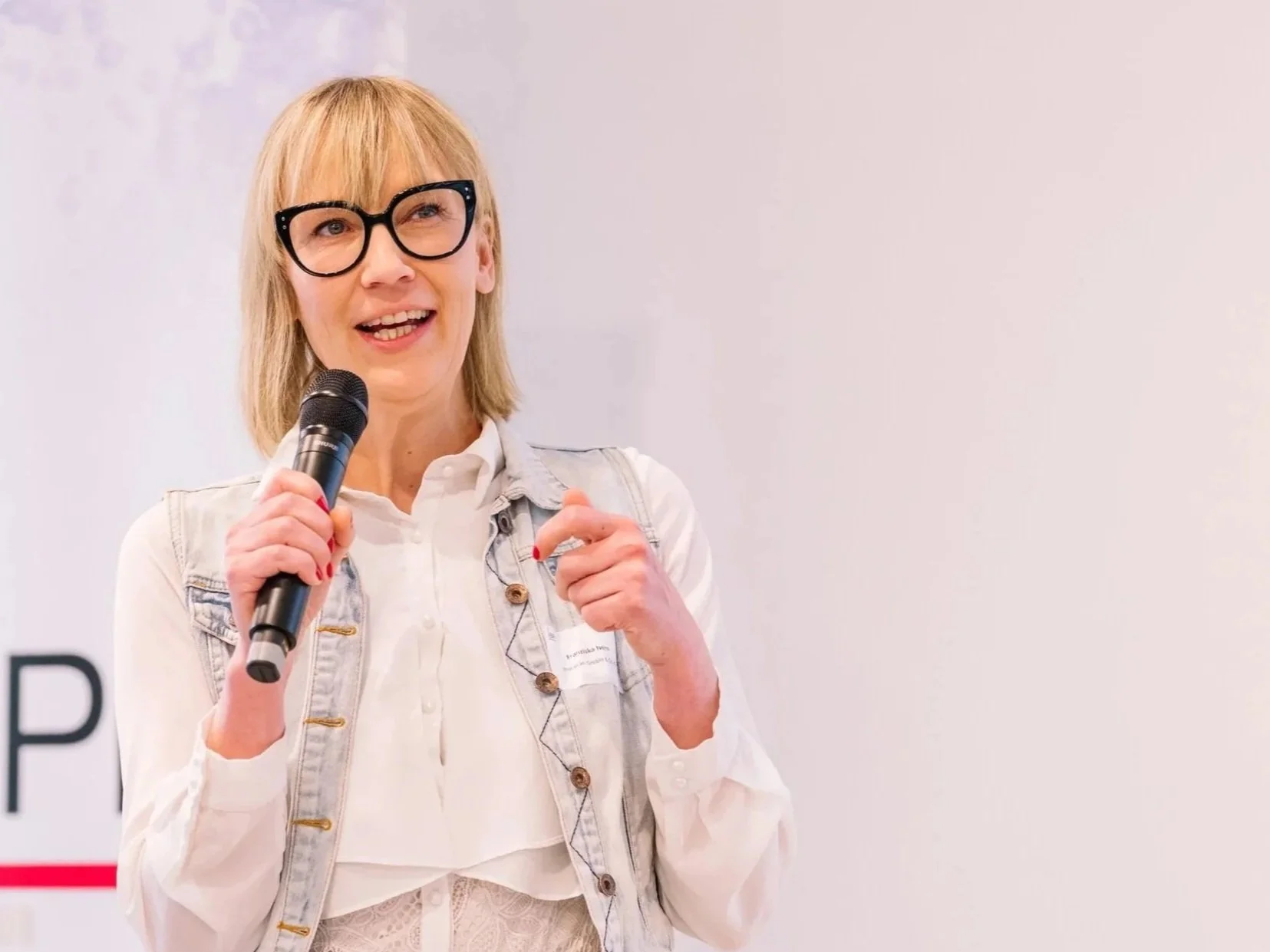 A woman with blonde hair, glasses, and a denim vest speaking into a microphone at an event.