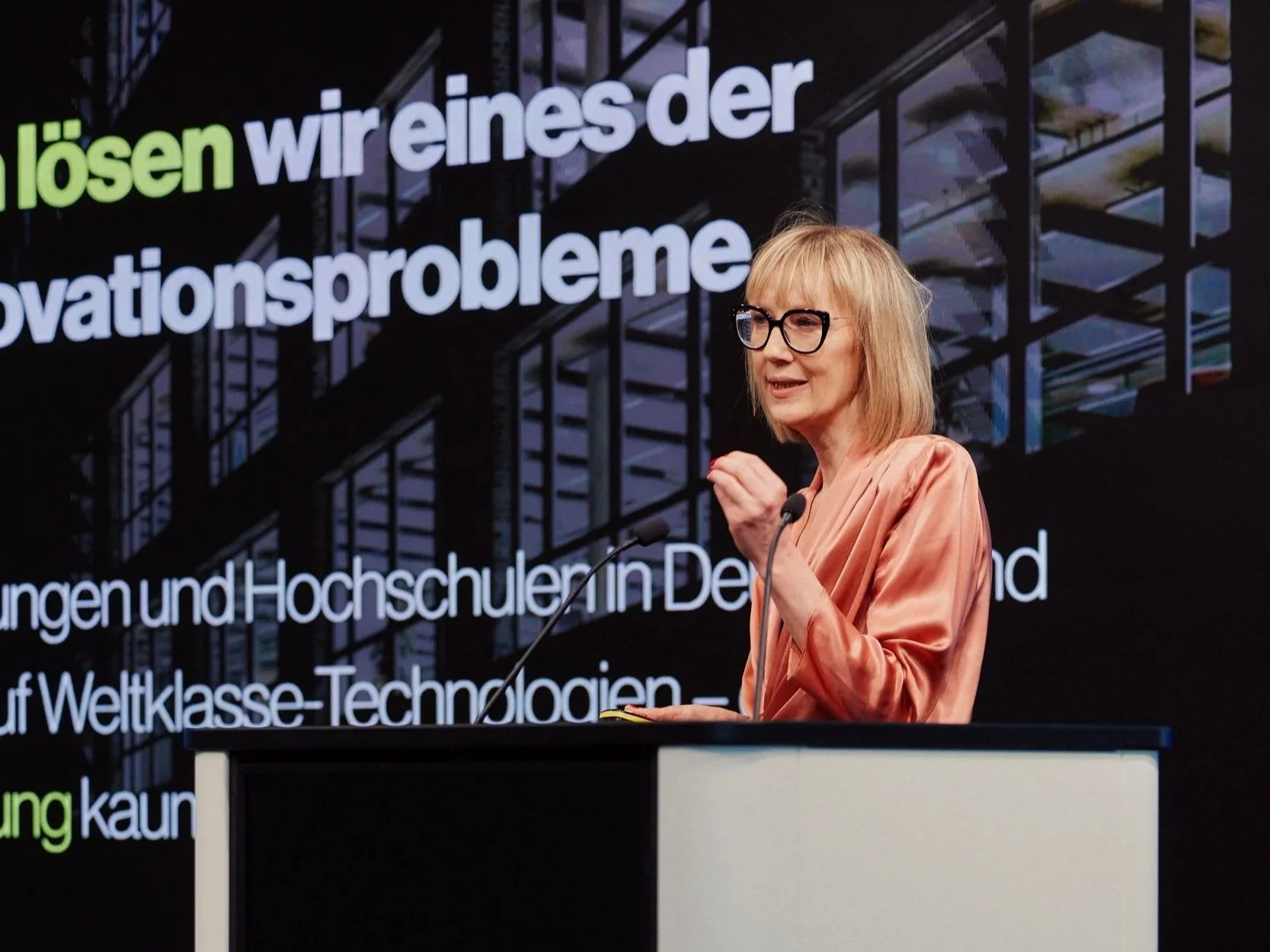 Eine Frau mit Brille und einem orangefarbenen Kleid spricht an einem Rednerpult bei einer Veranstaltung. Im Hintergrund ist ein großer Bildschirm mit deutschen Texten zu sehen.