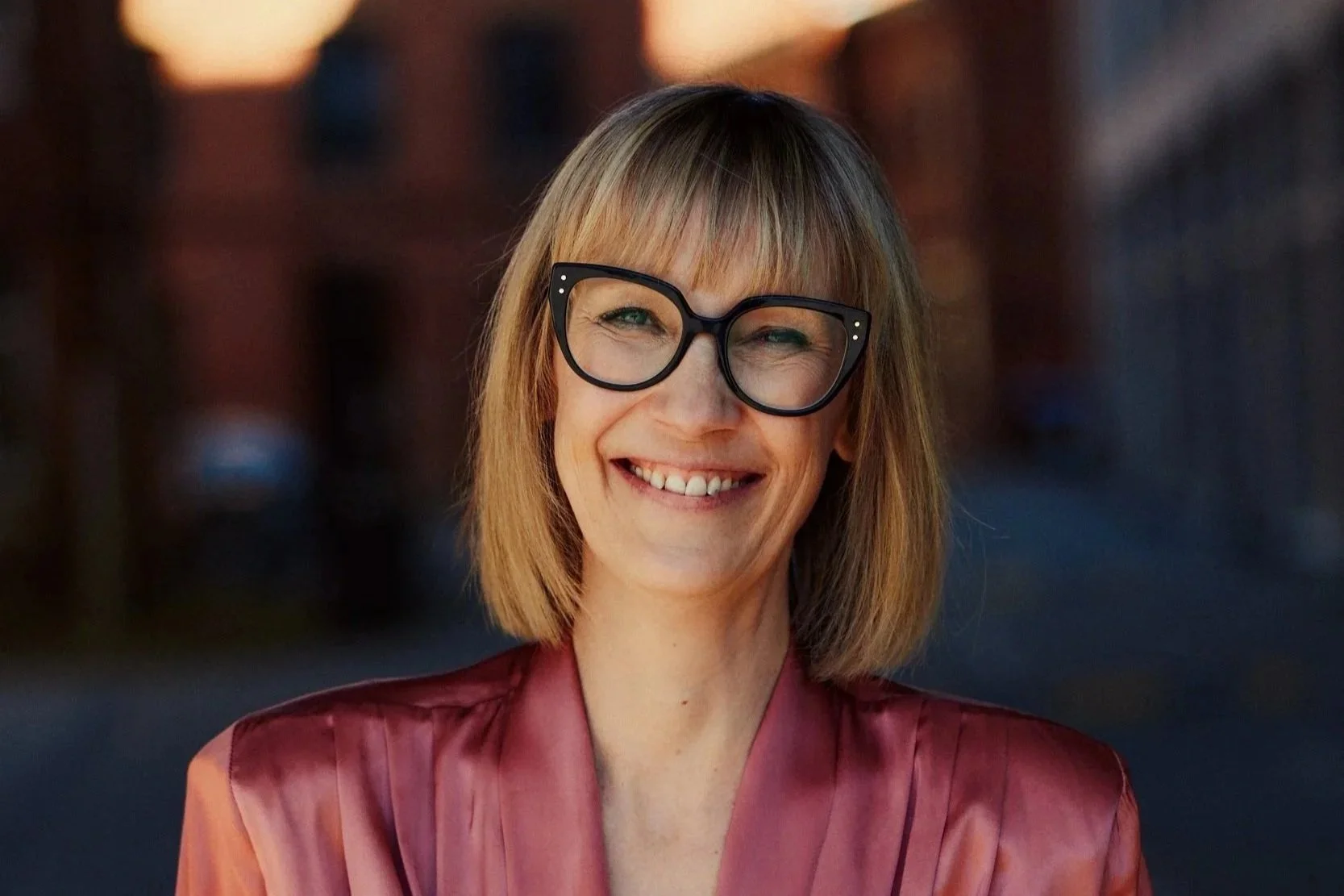 Eine lachende Frau mit Brille und geradem, kinnlangem Haar trägt ein rotes Outfit, steht vor einem unscharfen Hintergrund.