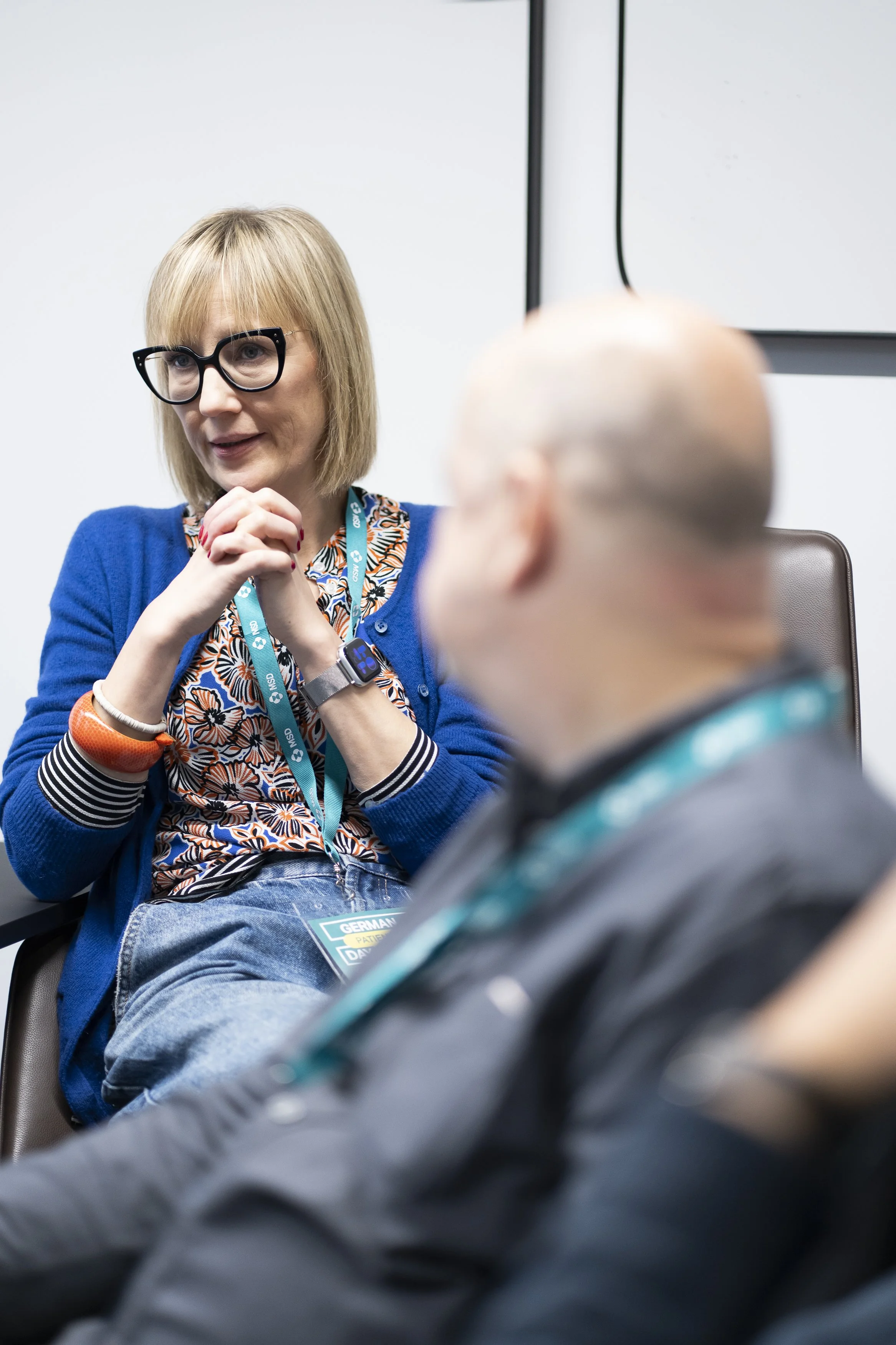 A woman with blonde hair, glasses, and a blue cardigan, talking to a bald man with glasses at an indoor conference or meeting.