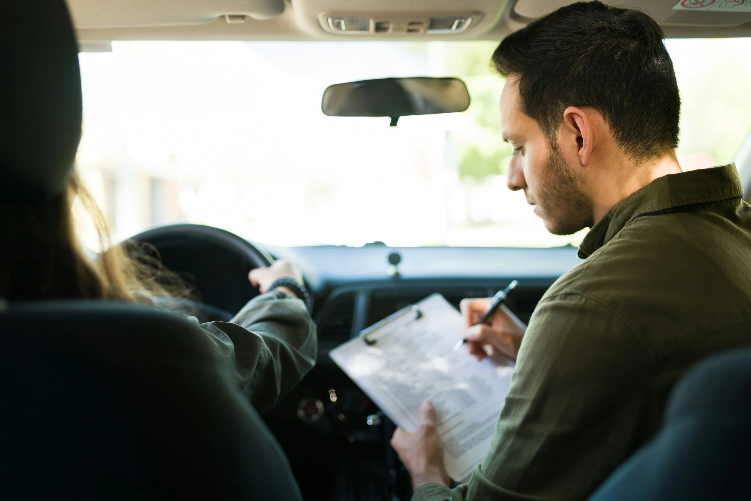 Two people sitting in a car; the driver is a woman, and the passenger is a man. The man is holding a clipboard and writing on it, while the woman is reaching the steering wheel. It appears to be daytime outside the car.
