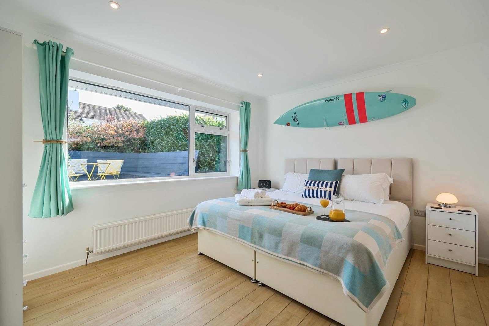 Bright bedroom with large window, white walls, beige headboard, and a surfboard mounted above the bed. Bed has white and blue linens, with a blue pillow and breakfast tray with orange juice and pastries. White bedside table with a small lamp and a gl