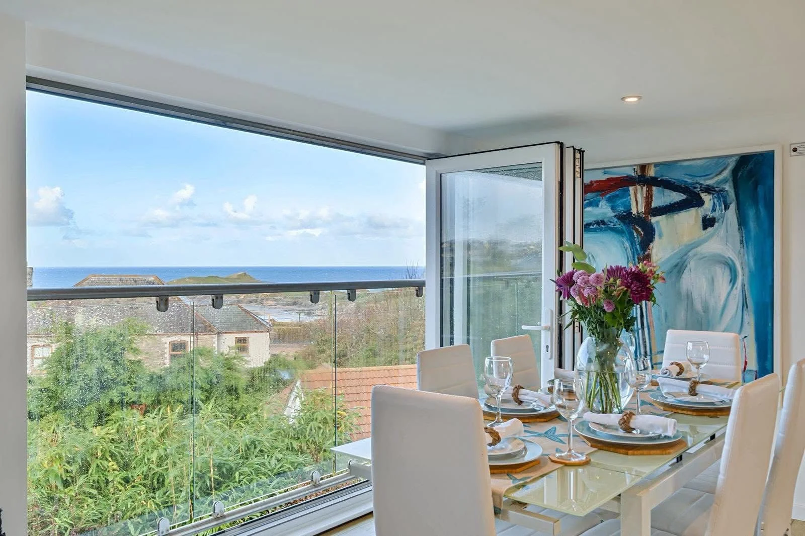 A dining room with a large glass window showing a view of the ocean and a partly cloudy sky. The room is decorated with a vase of flowers on the table, which is set with plates, wine glasses, and napkins. There is a colorful abstract painting on the 
