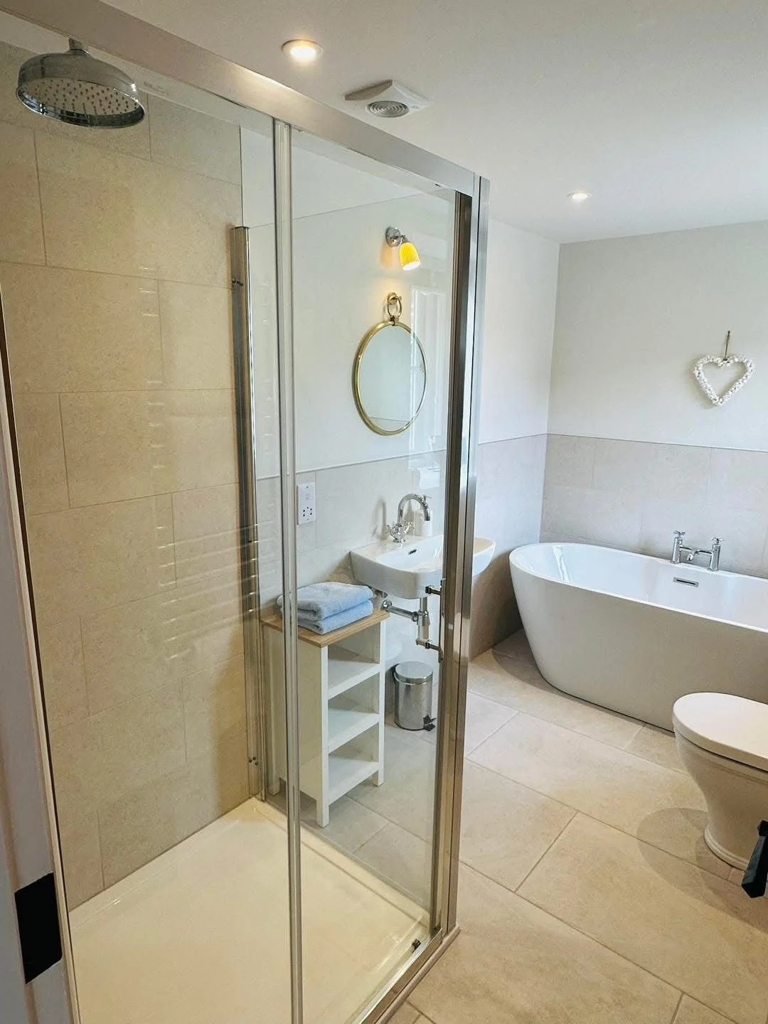 Modern bathroom with a glass shower enclosure, a freestanding bathtub, a toilet, and a small white sink with a round mirror above. Decorations include a heart-shaped wall hanging and a towel on a small wooden shelf.
