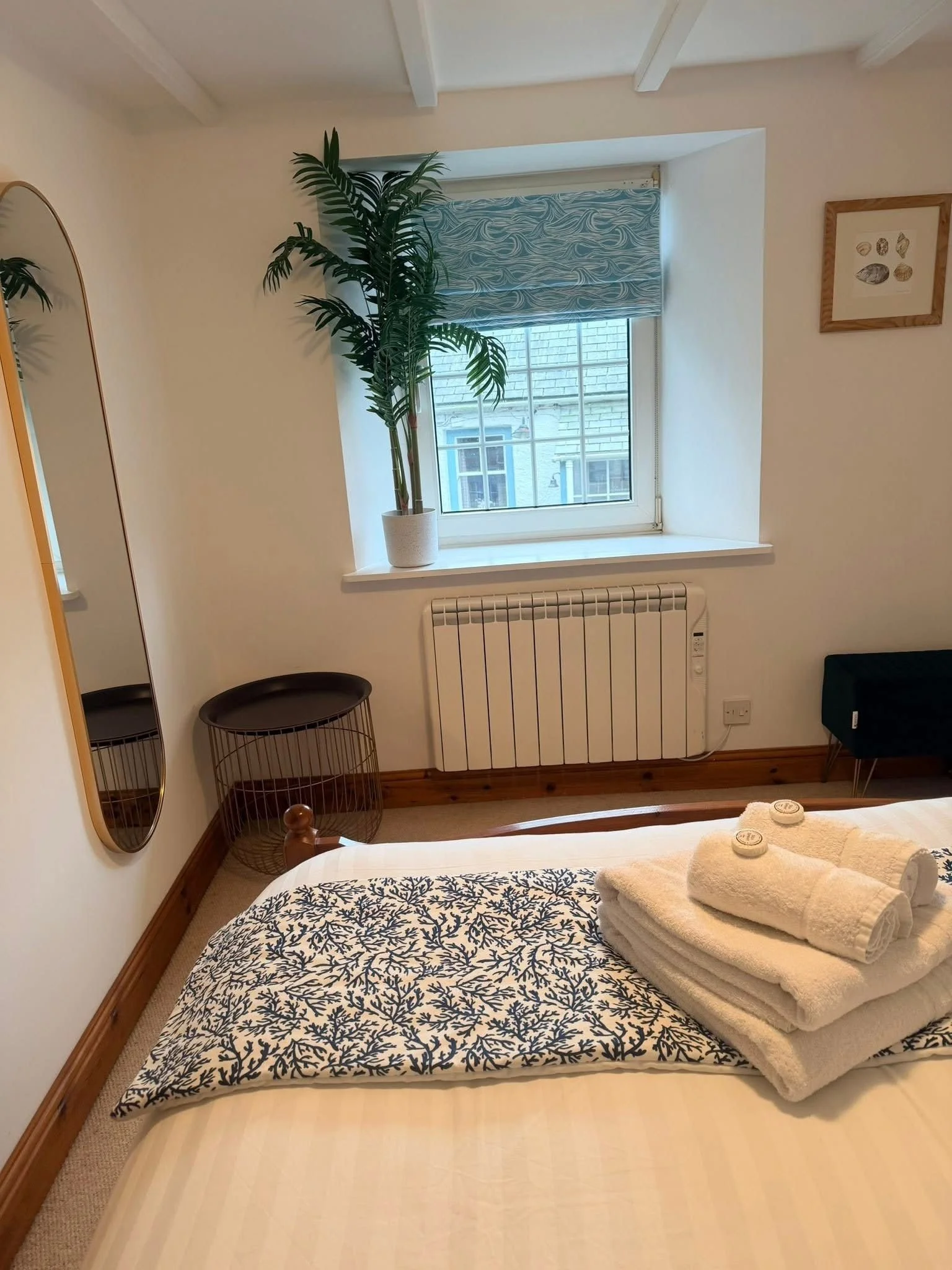 A cozy bedroom with a bed covered in a navy and white coral-patterned bedspread. On the bed are neatly folded white towels. To the left of the bed, there's a round wire basket and a wall mirror. The room has a window with a blue patterned curtain, a 