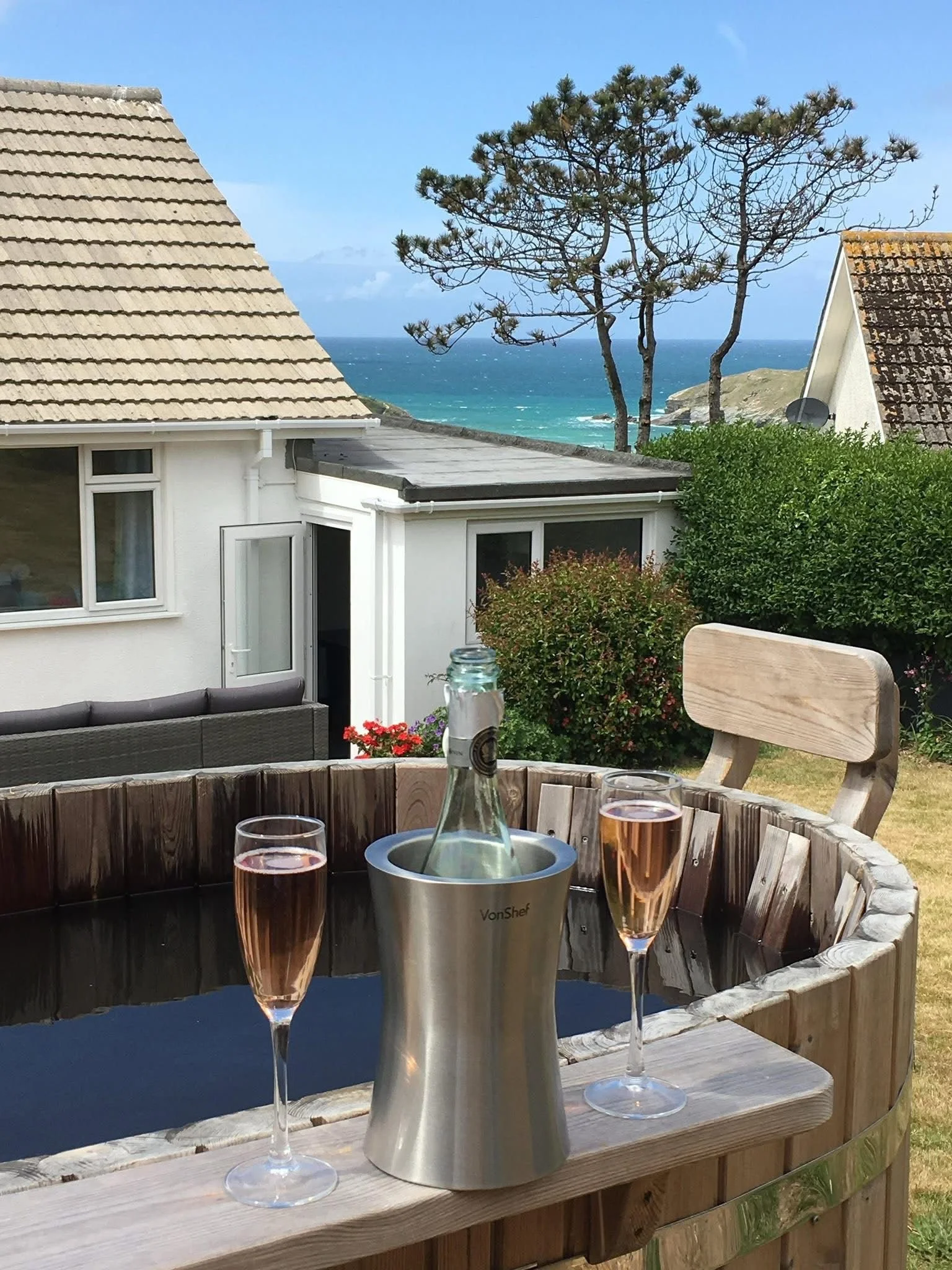 Three glasses of rosé wine, a champagne bucket with a bottle, on a wooden table outdoors overlooking the ocean with houses, trees, and a rocky coastline against a blue sky.