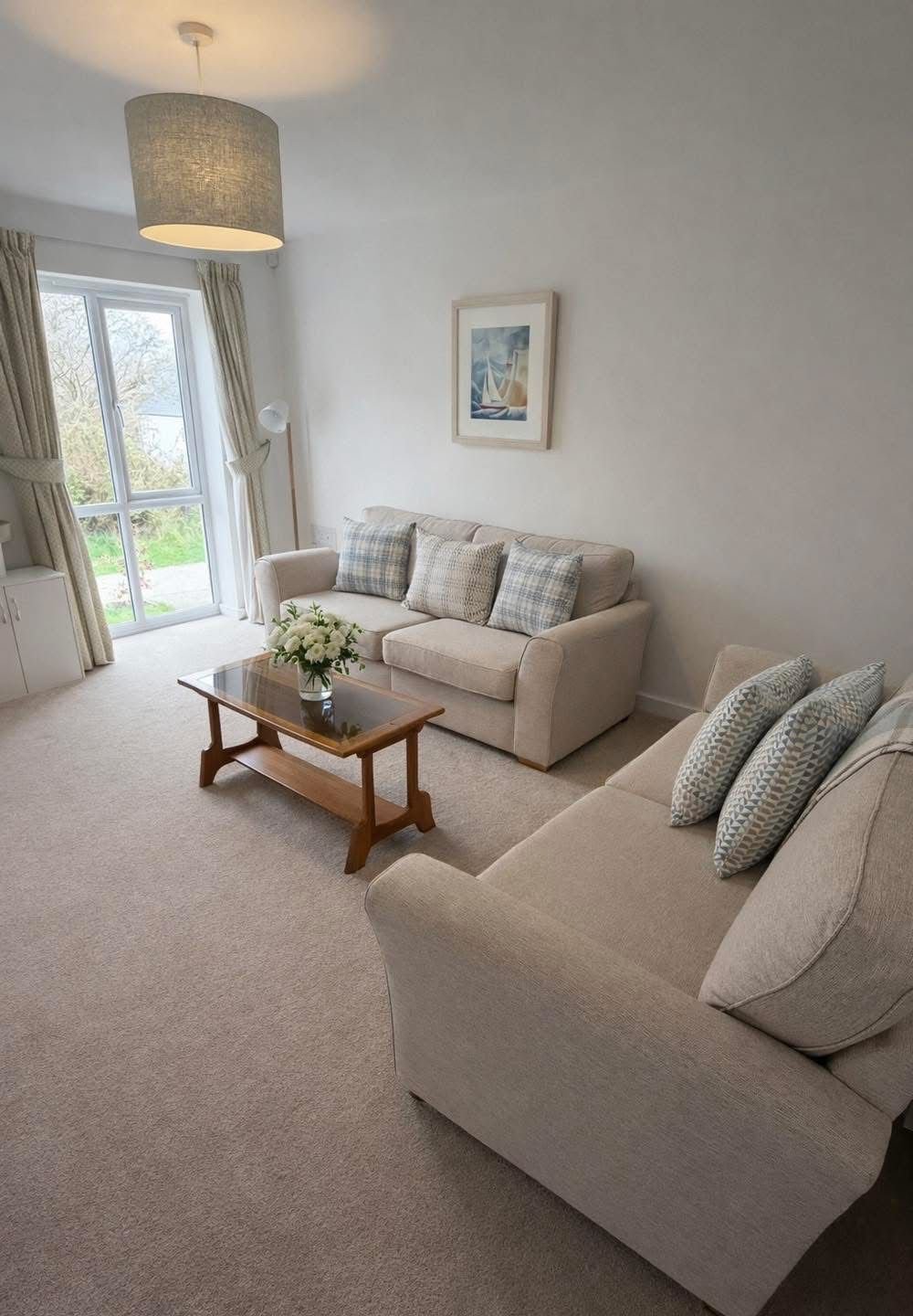 Bright living room with two cream-colored sofas with plaid cushions, a wooden coffee table with a flower vase, a large window with beige curtains, and a ceiling lamp. A framed picture hangs on the wall.