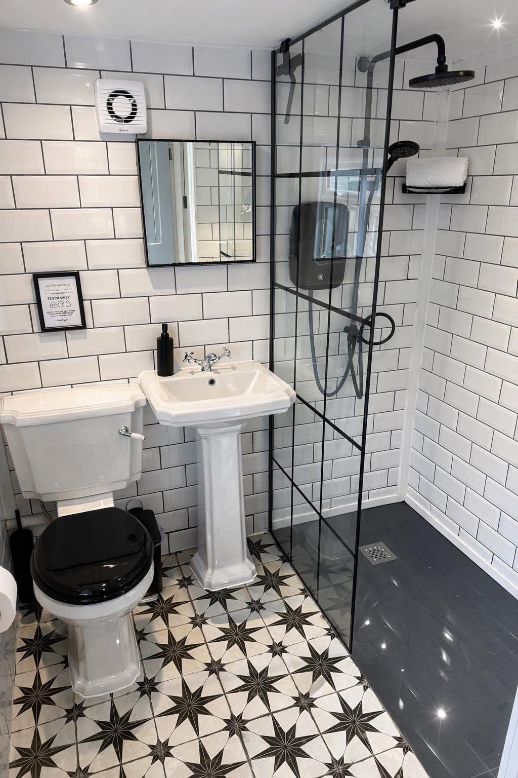 A modern bathroom with black and white geometric floor tiles, white subway wall tiles, a small white pedestal sink, a black toilet seat, a mirror, a shower area with a black-framed glass partition, black shower fixtures, a wall-mounted soap dispenser
