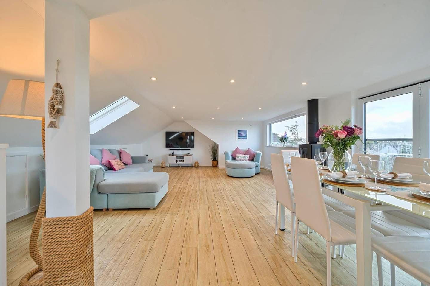 Bright, airy living and dining room with hardwood floors, blue and pink accents, a flat-screen TV, a fireplace, and large windows.