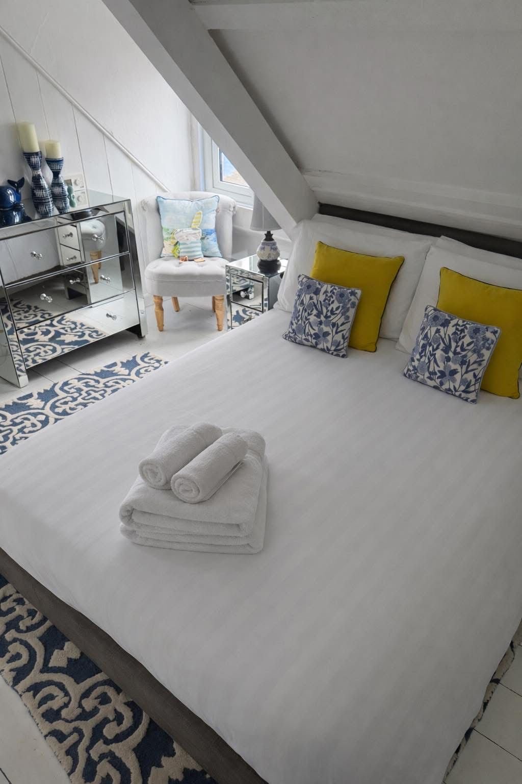 A neatly made bed with white linens and pillows, including yellow and blue patterned pillows, with towels folded on top. The room has a patterned rug, a small mirrored dresser with decorative candles and a teapot, a white upholstered armchair with a 