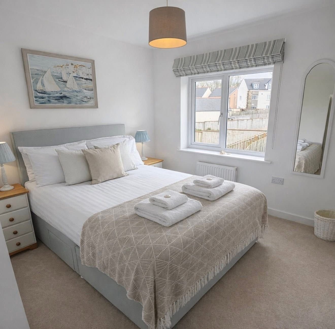 A neatly made bedroom with a double bed, beige and white bedding, and pillows. Two nightstands with lamps are on either side. There are folded towels on the bed. A window with a striped valance shows a view of neighboring houses and a wooden fence. A