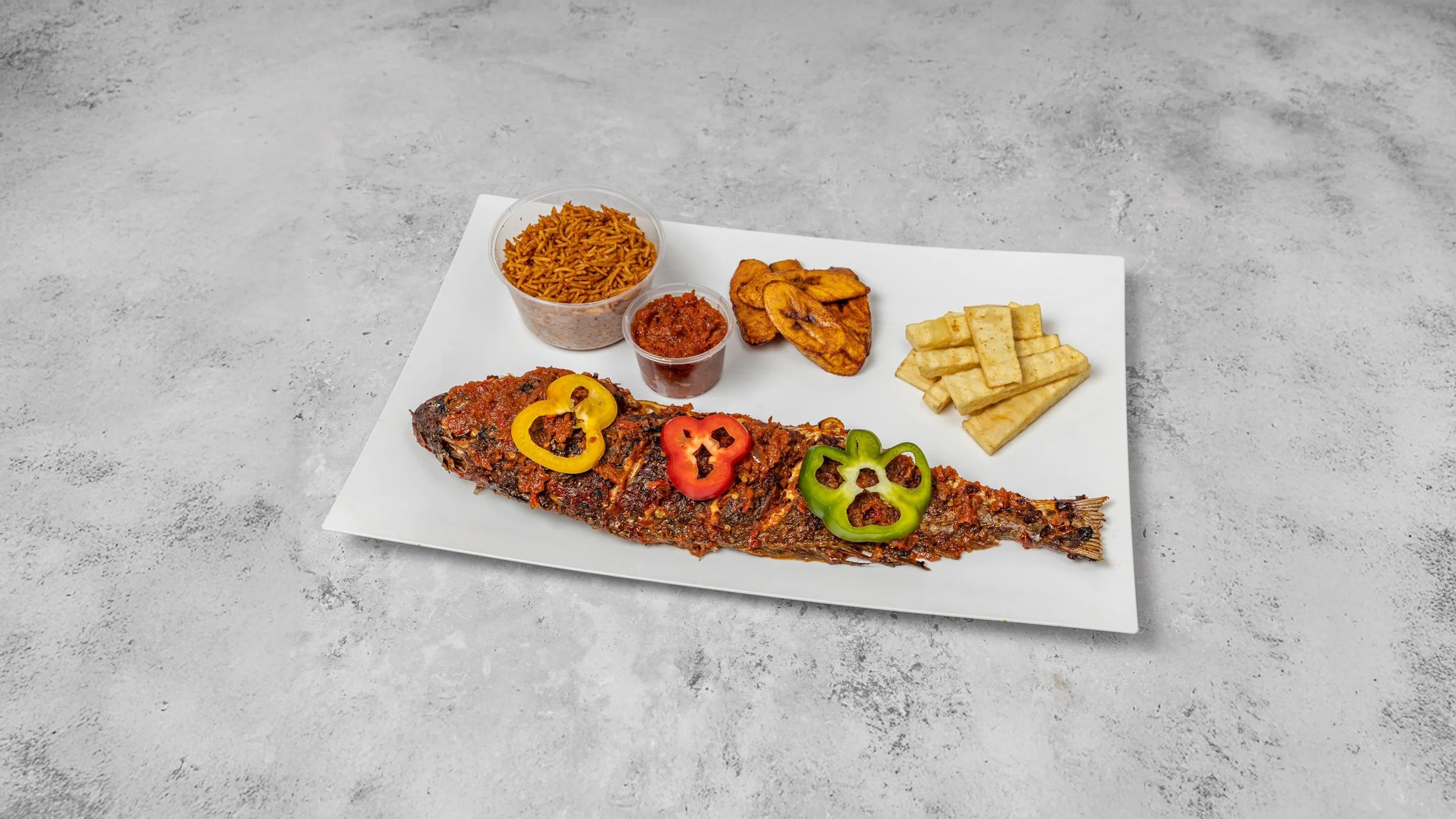 Whole grilled fish topped with sauce and garnished with red, yellow, and green bell pepper slices on a white plate, accompanied by rice, plantains, French fries, and two small cups of sauce.