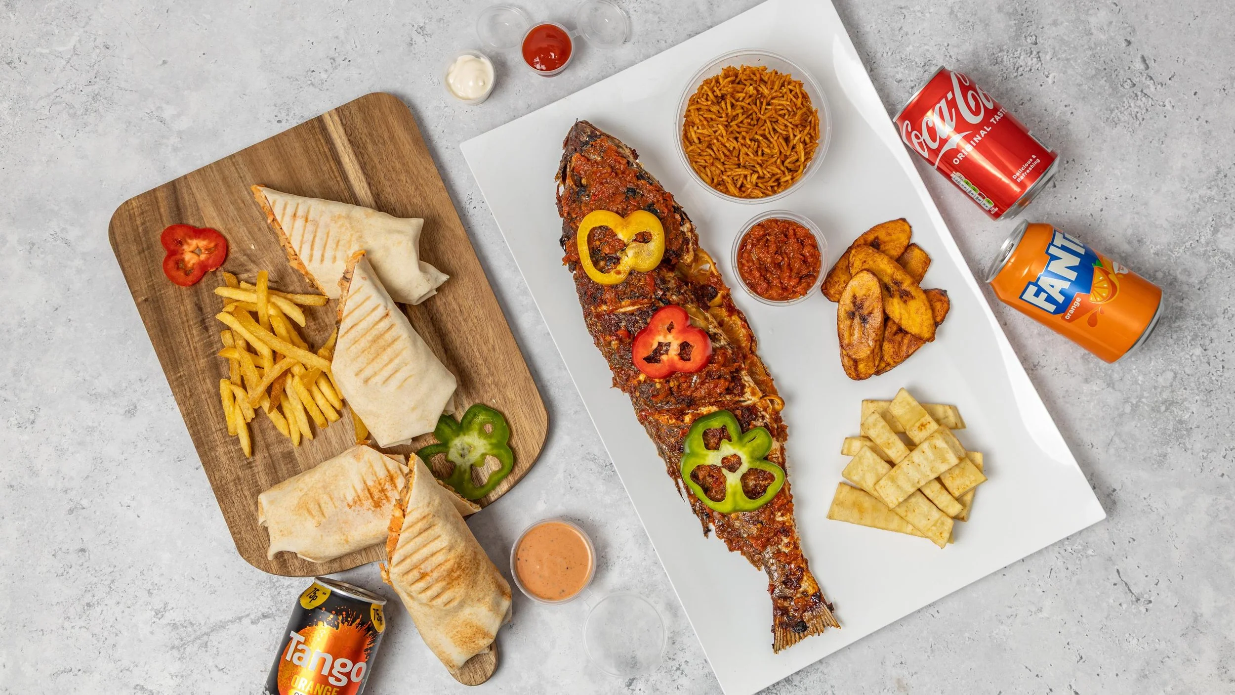 A meal with a grilled fish covered in spicy sauce and garnished with sliced green and red peppers, surrounded by sides of French fries, potato chips, and tortillas filled with fries, along with small cups of ketchup, mayonnaise, and spicy sauce, and 