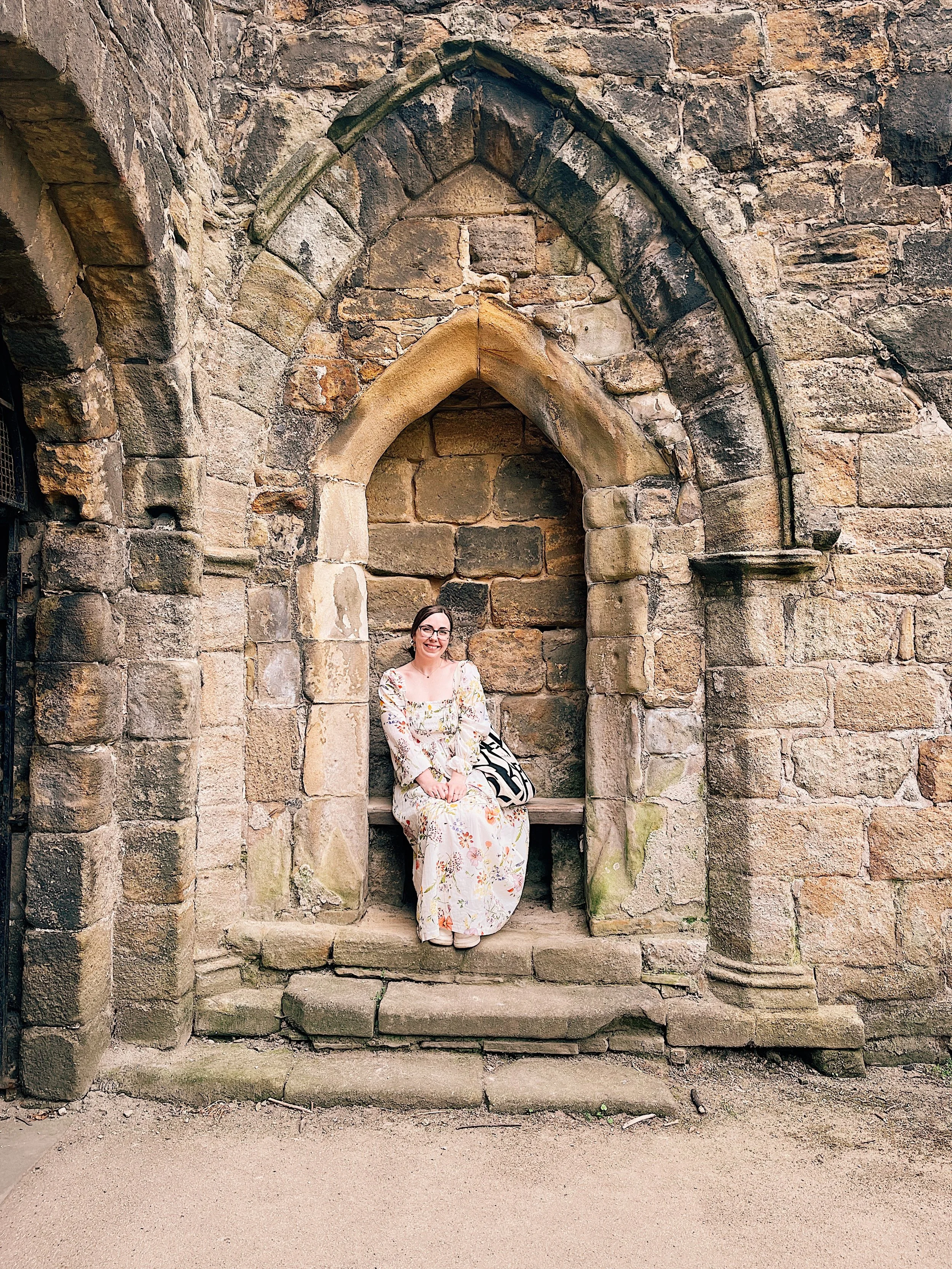 A woman in a floral dress sitting on a stone bench inside a small alcove with a pointed arch in an old stone wall.