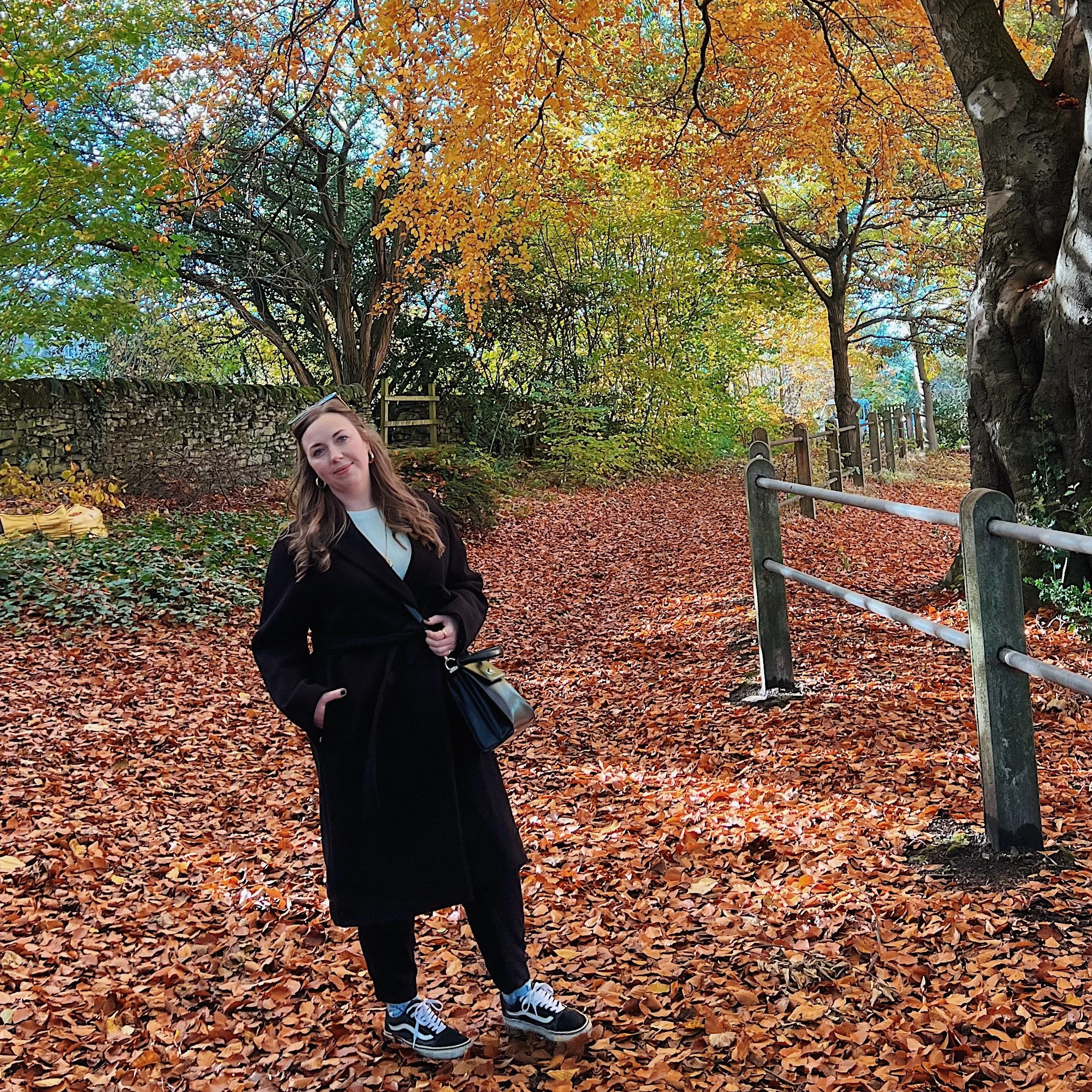 Young woman standing on a leaf-covered path in a park during autumn, with colorful orange, yellow, and green trees and a stone wall in the background.