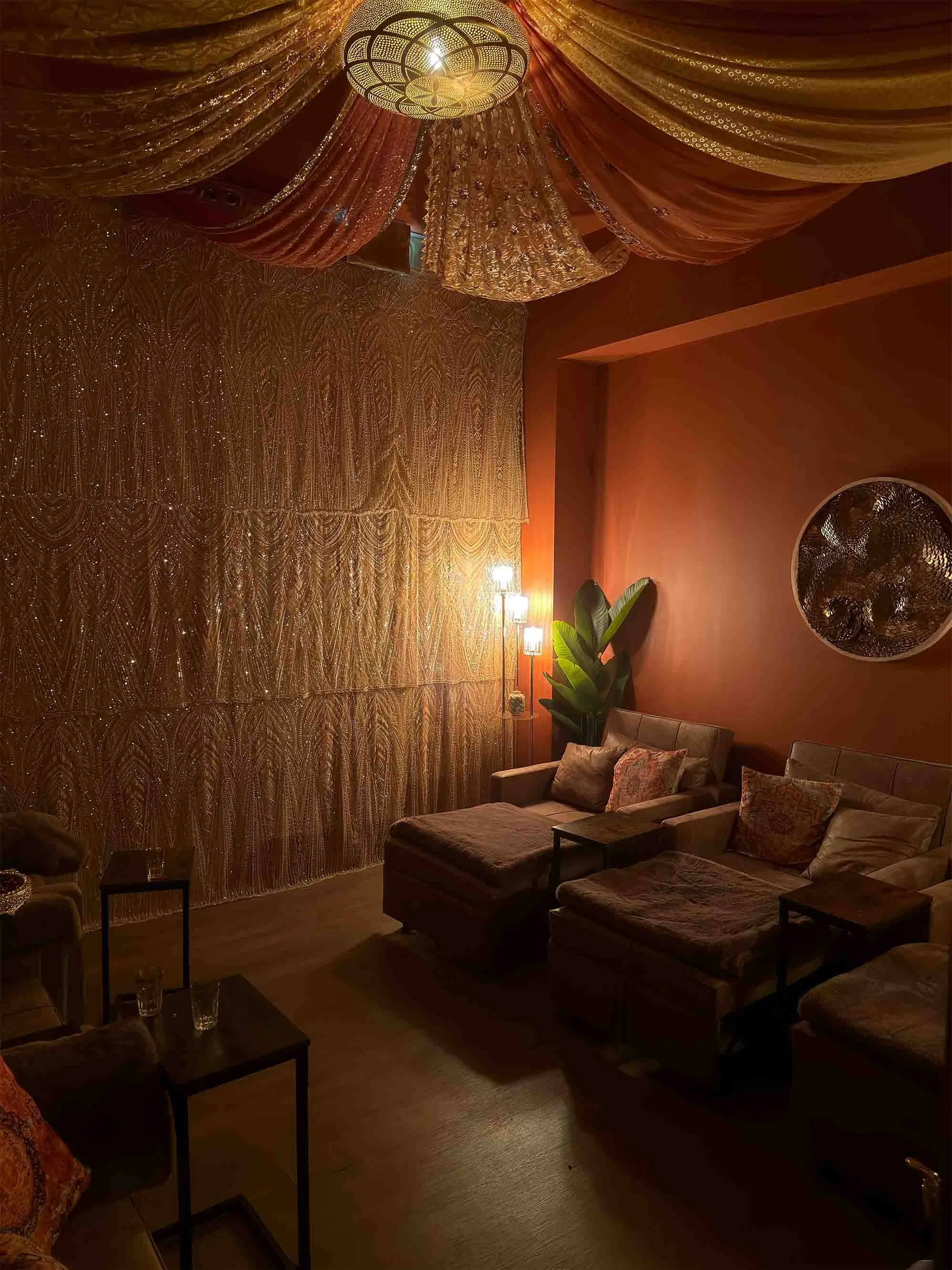 A cozy, dimly lit room with orange walls, adorned with gold and orange draped fabric on the ceiling, gold curtained wall, and modern wall art. There are plush sofas with cushions, small tables with glasses, and a large leafy plant, illuminated by a standing lamp and decorative ceiling light.