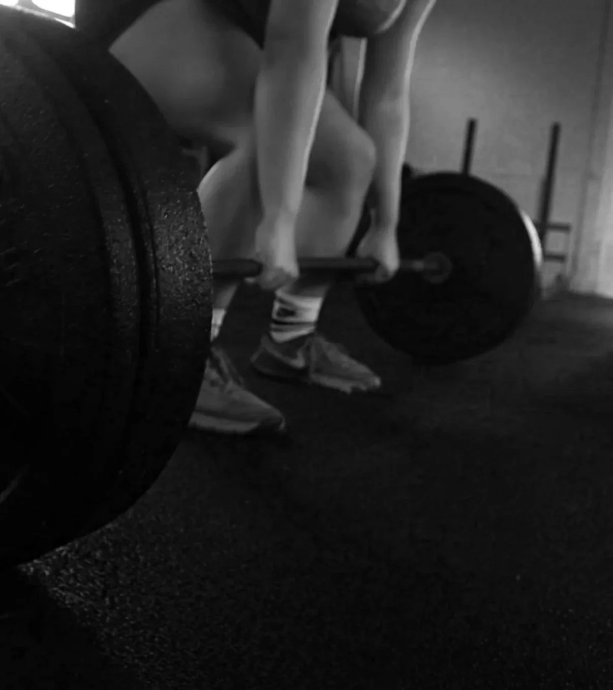 A person standing behind a barbell with weights in a gym, seen from a low angle in black and white.