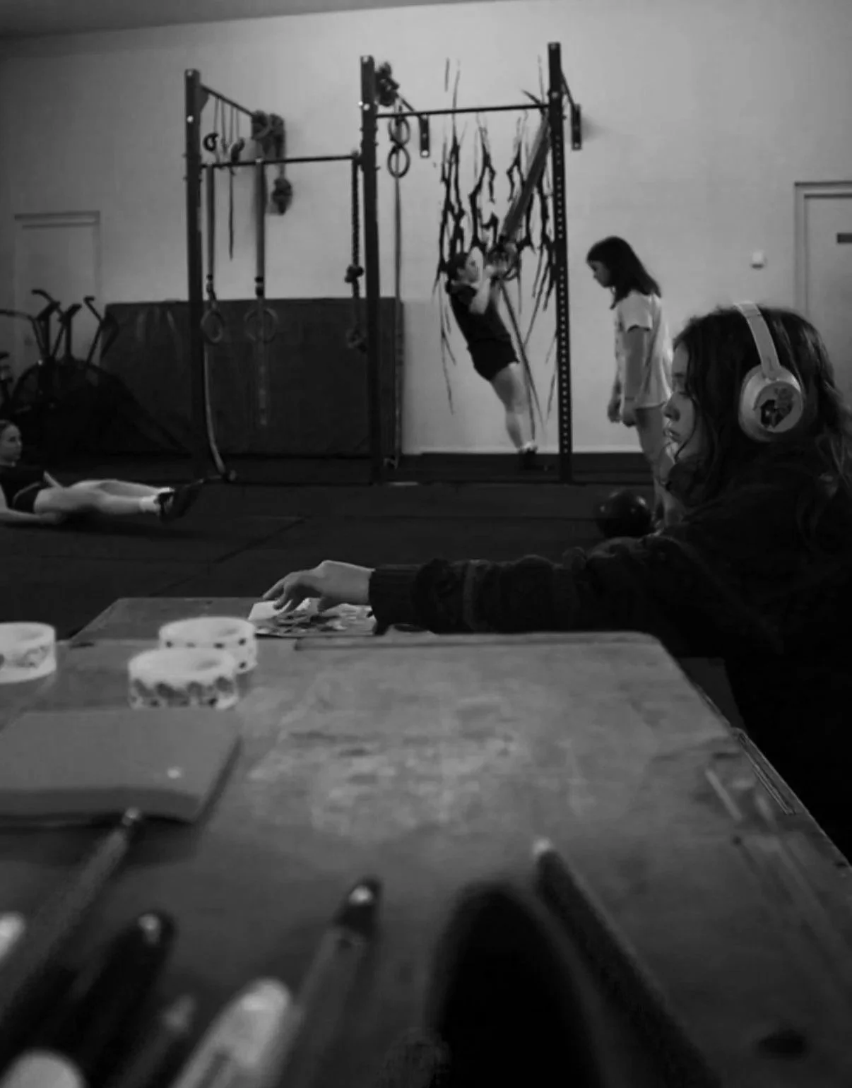 A girl practicing gymnastics on rings in a gym, with two other girls watching nearby. A woman wearing headphones is sitting at a table in the foreground, looking at playing cards or photos, and there are notebooks and writing utensils on the table.
