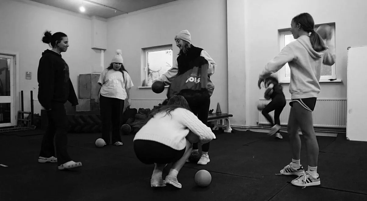 Group of young girls and an adult woman in a gym, engaging in fitness activities with medicine balls and jumping exercises.
