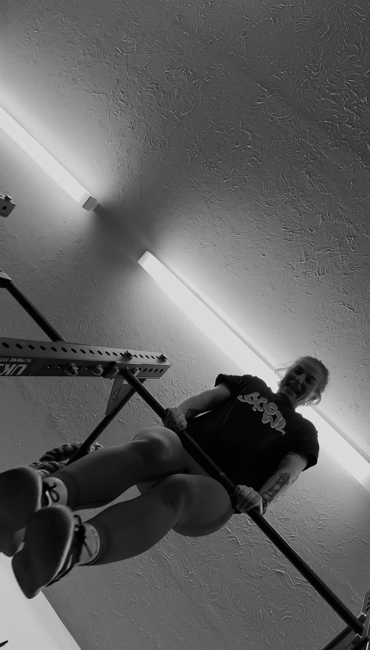 A woman performing a pull-up exercise on a bar in a gym, with a textured ceiling and fluorescent lights visible overhead.