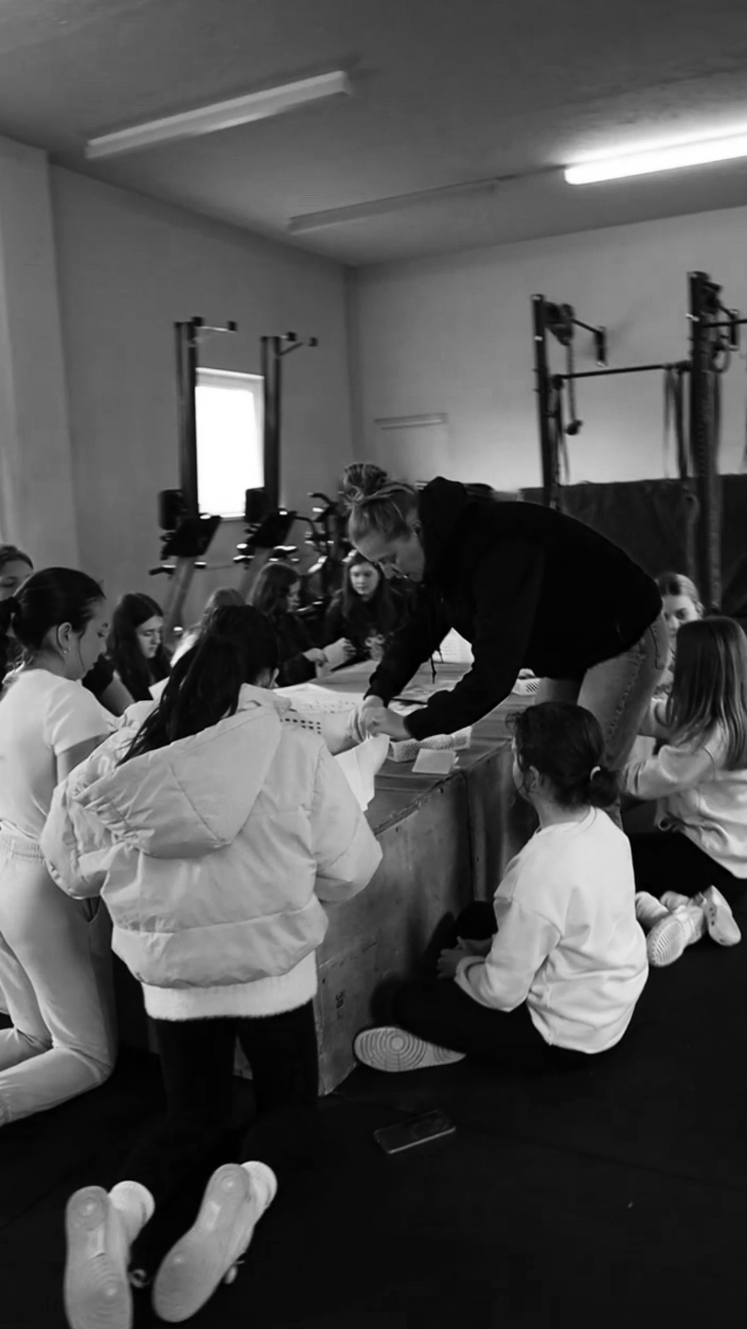 A group of girls in a gym, some seated and others kneeling, receiving instructions from a woman standing at a table. The gym has workout equipment like rowing machines and pull-up bars in the background.