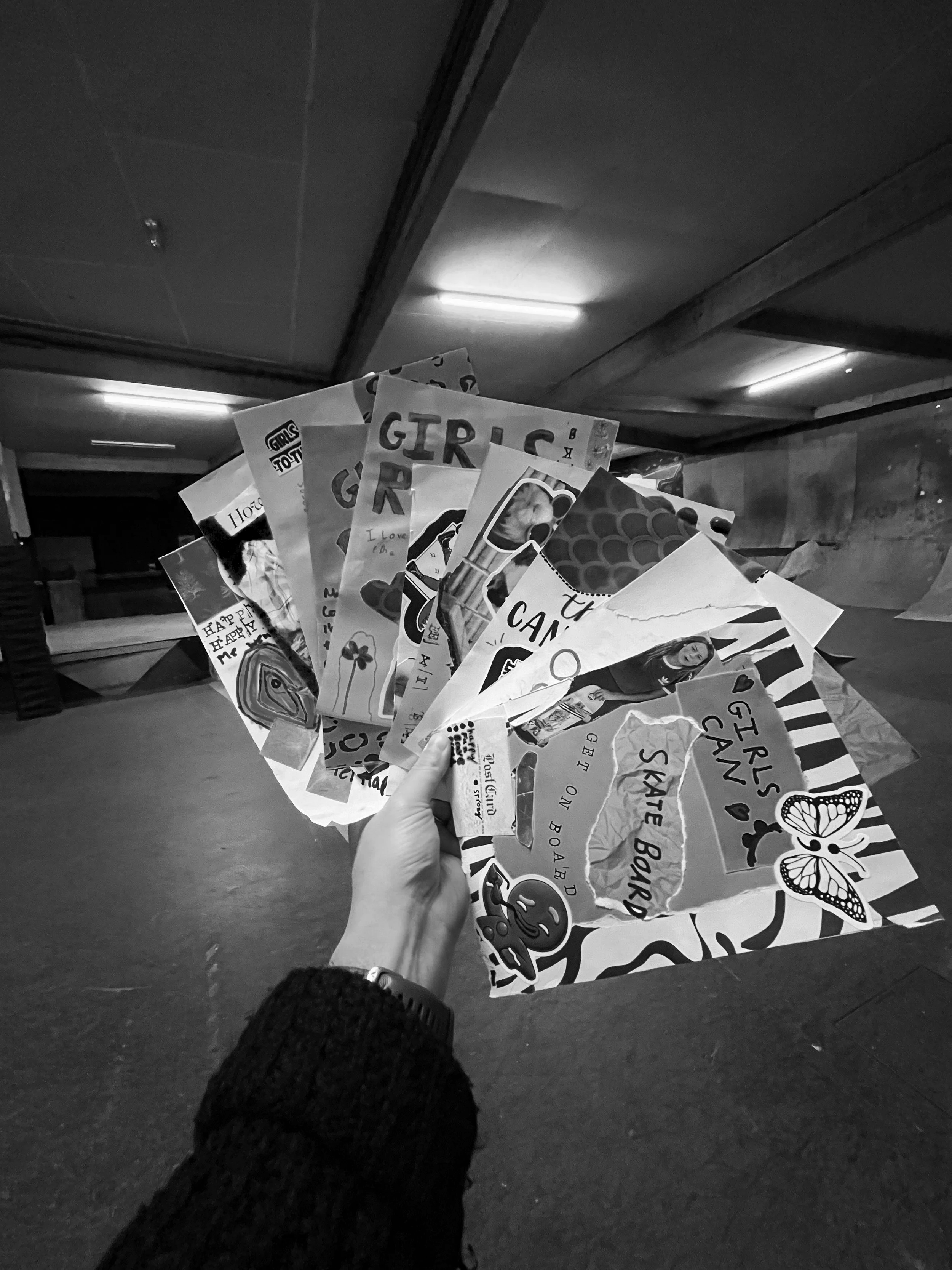 Hand holding several colorful, patterned paper flyers or posters in a dark indoor skate park or recreational area.