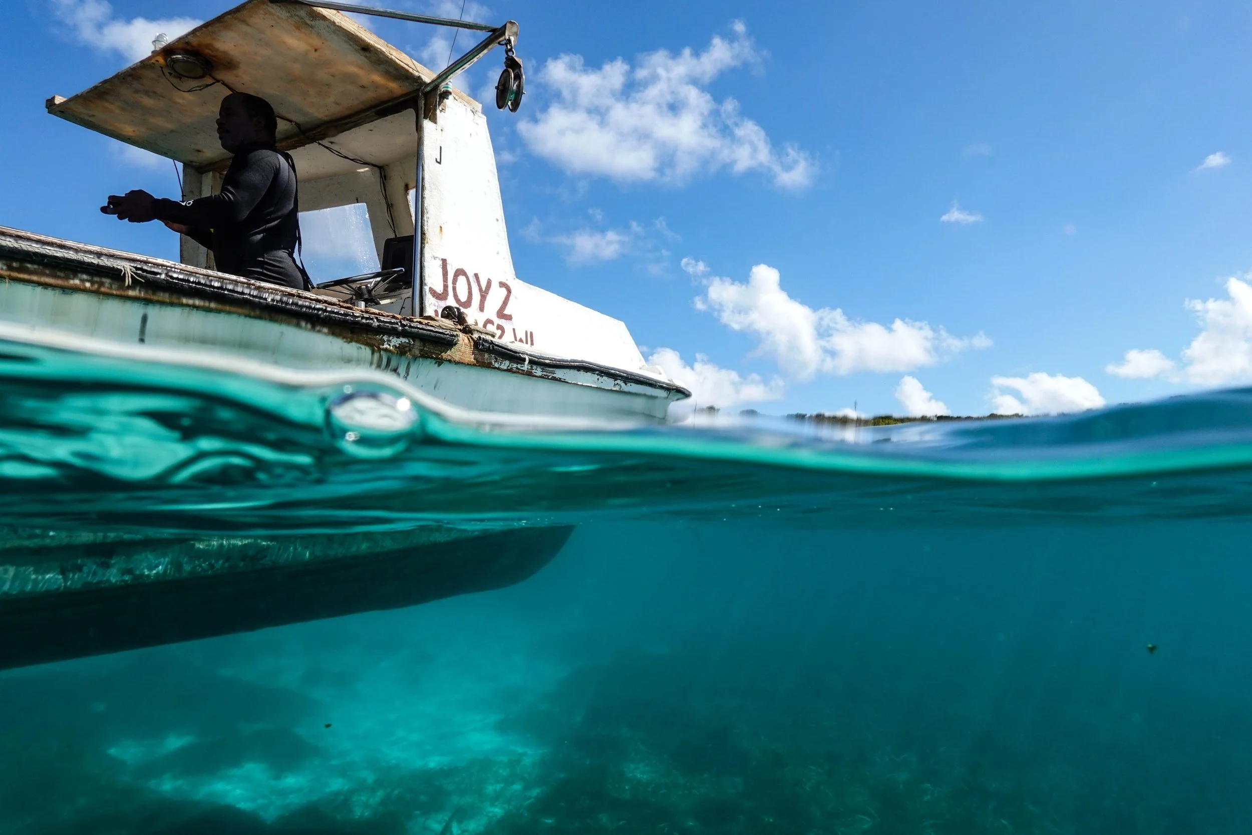 Half underwater, half above water view of a boat with a person on board, blue sky with clouds in the background.