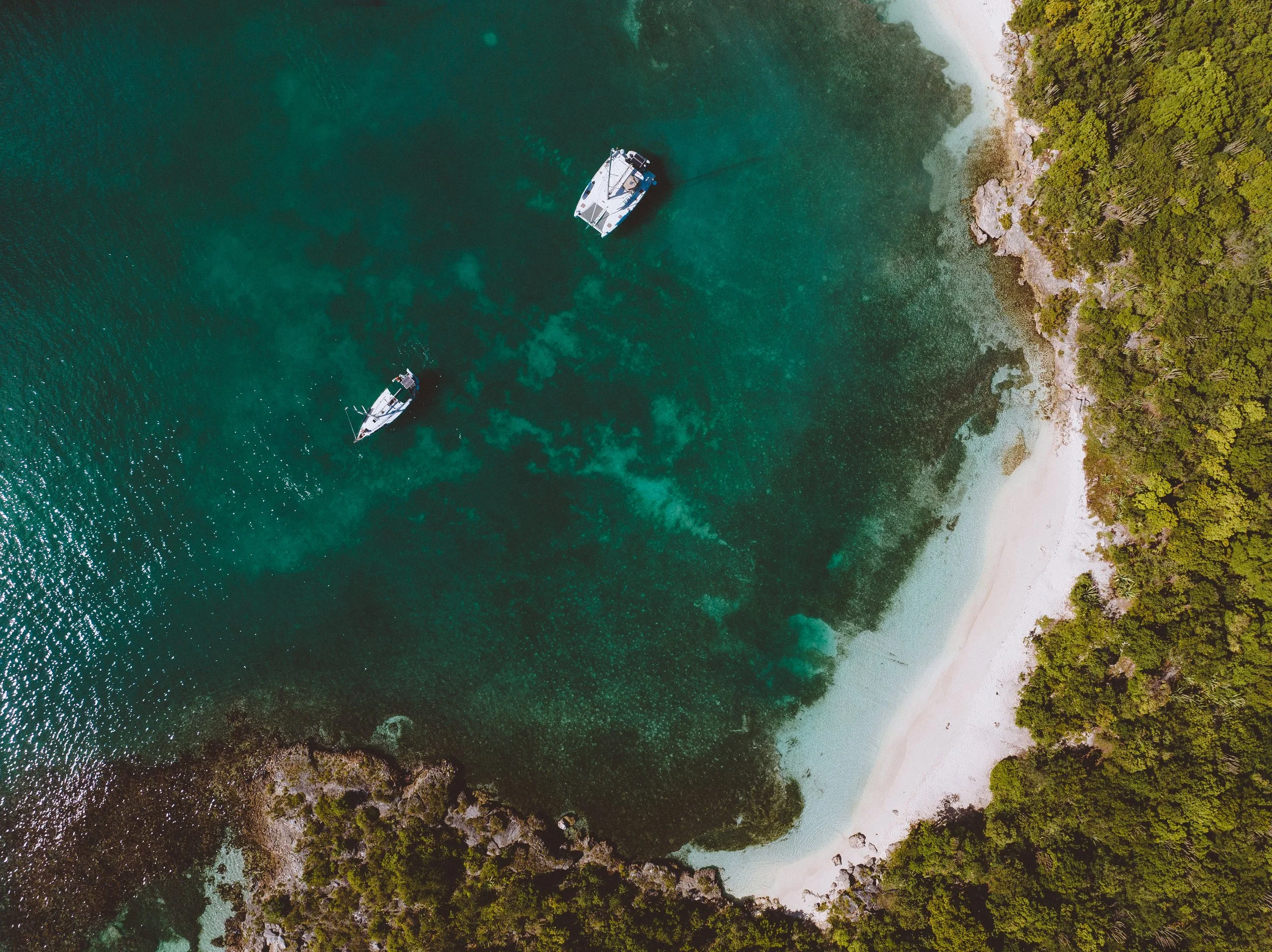 Aerial view of a shoreline with two boats floating in clear turquoise water surrounded by dense green trees.