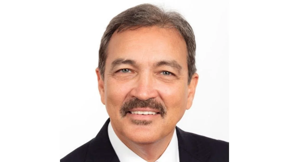 Headshot of a middle-aged man with brown hair, a mustache, wearing a black suit and white shirt, smiling against a white background.