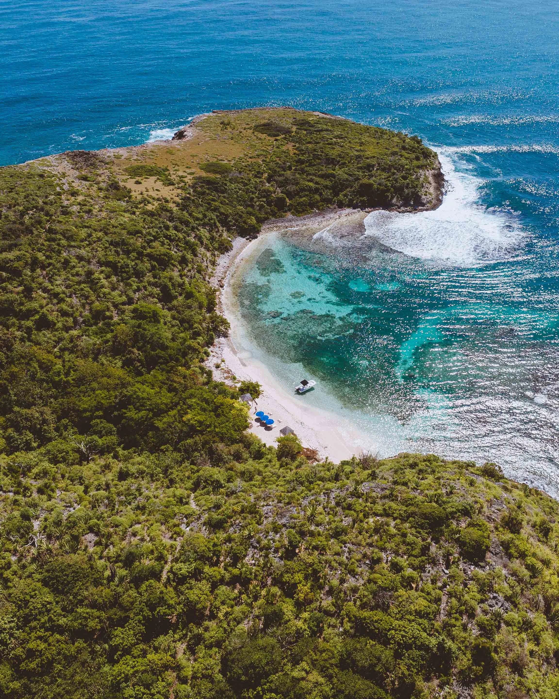 Aerial view of a small, curved beach surrounded by lush green vegetation and rocky cliffs, with clear blue water and a boat anchored near the shore.