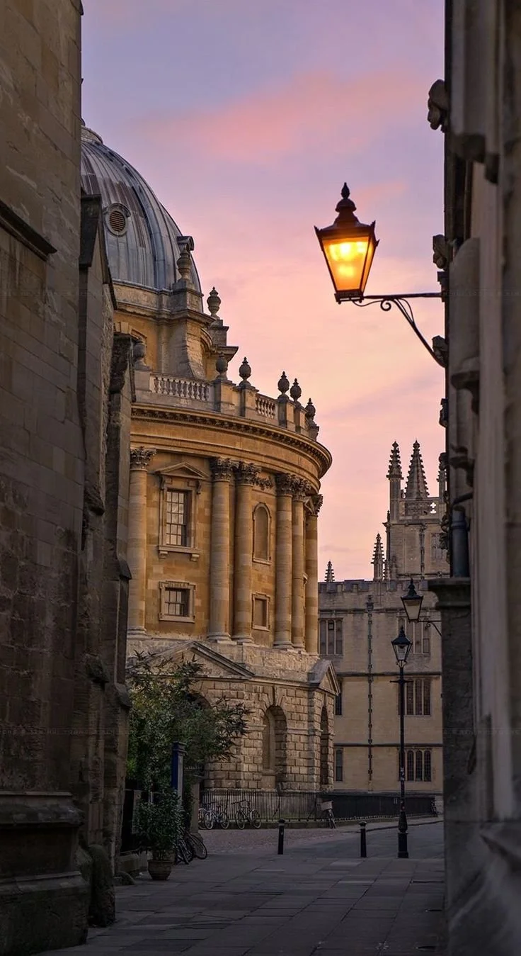 A historic European street scene at sunset with classical architecture, a domed building, Gothic spires, and a glowing street lamp against a pink and purple sky.