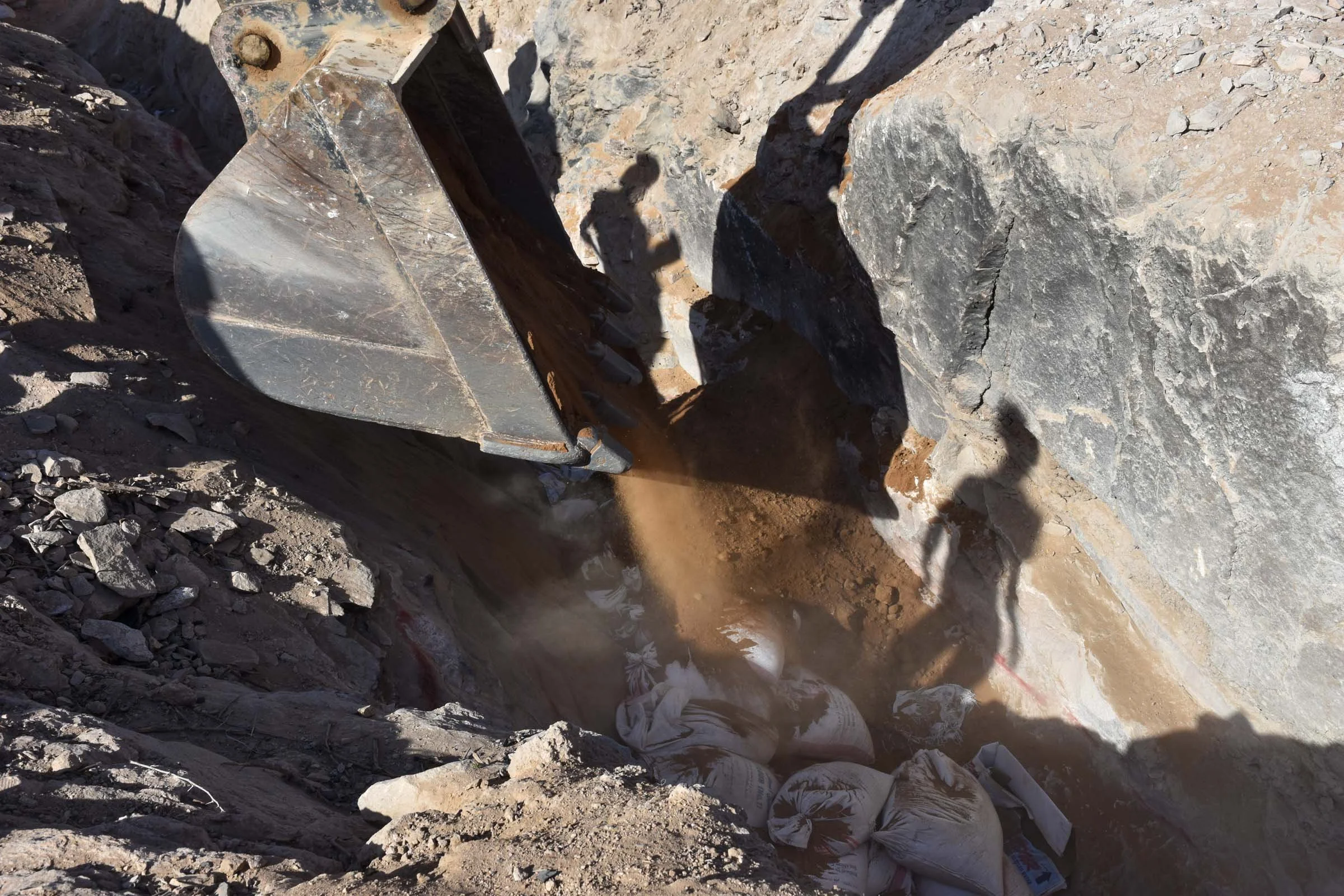 An excavator bucket tips earth into a ditch lined by people's shadows