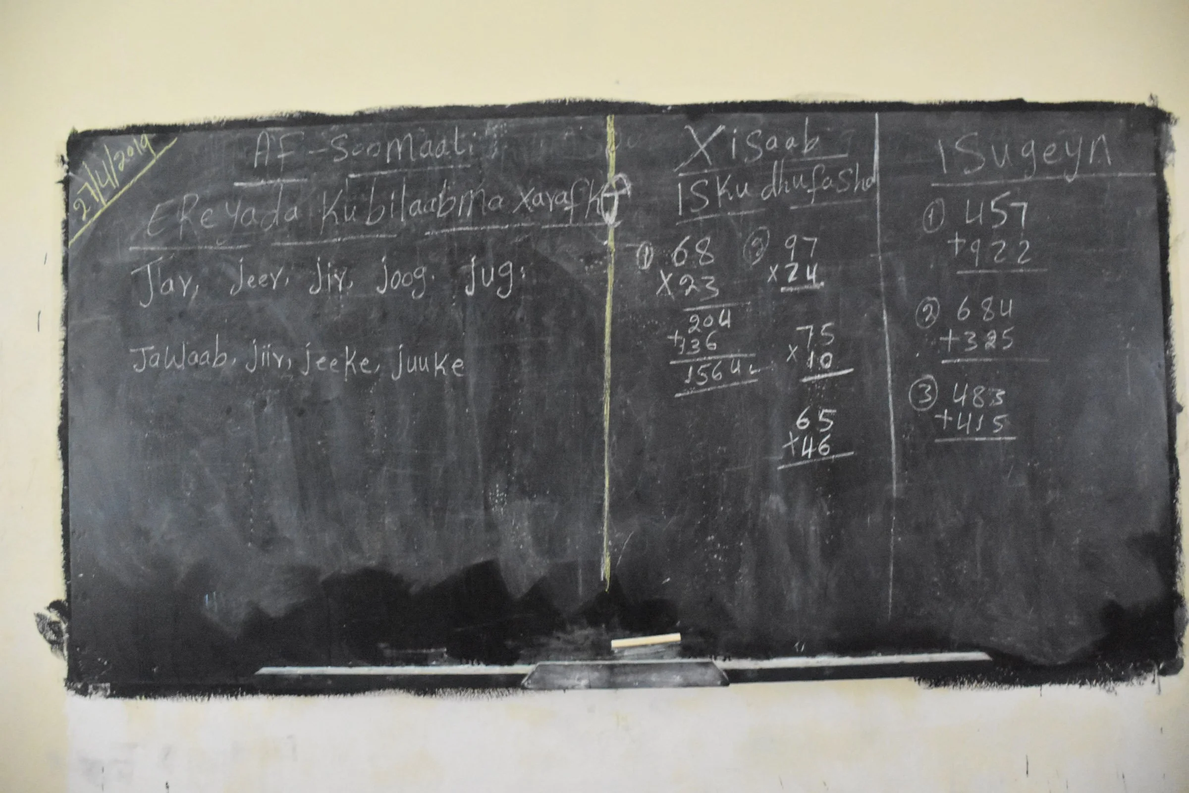 A blackboard shows Somali language, addition and multiplication exercises on the wall of a classroom