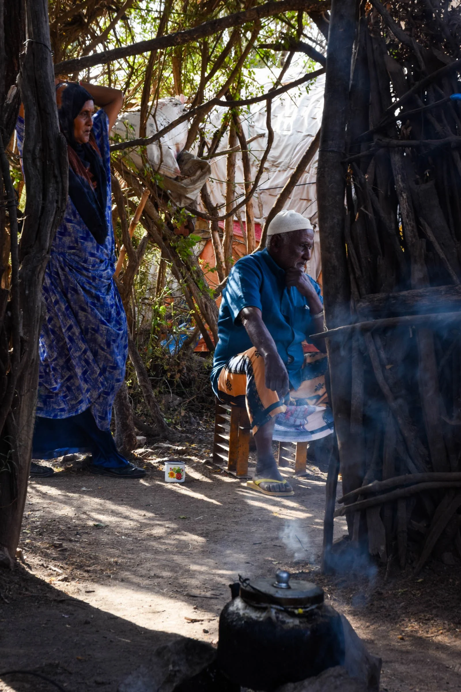 A black cooking pot spouts team in front of a man and woman seen through a wooden door frame