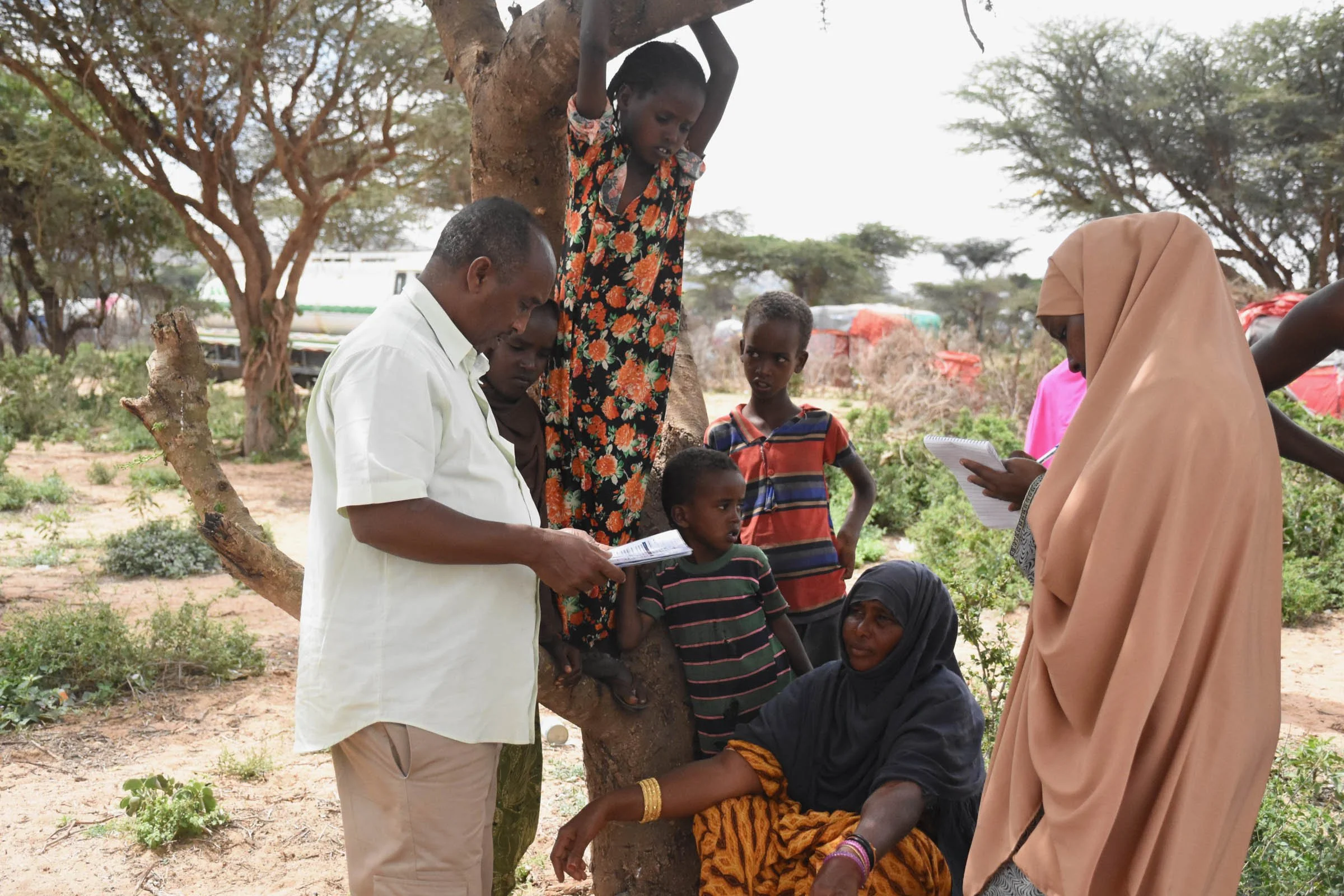 A girl hangs from a tree under which boys stand and a woman sits talking to a man and woman conducting a survey