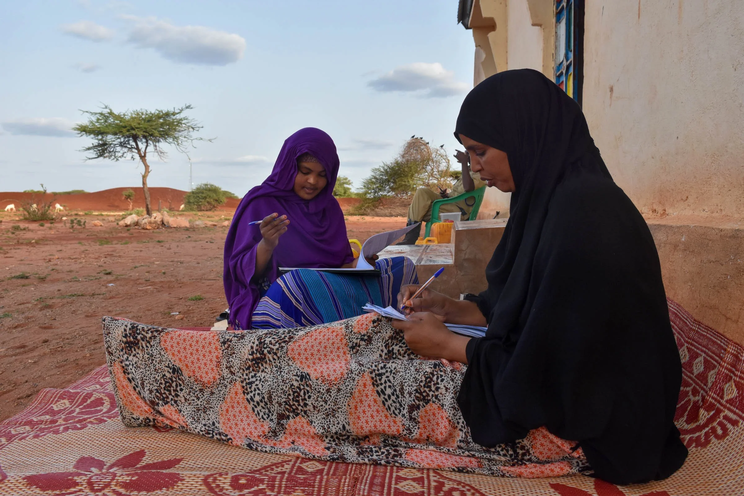 Two women sit on a mat on the ground while writing