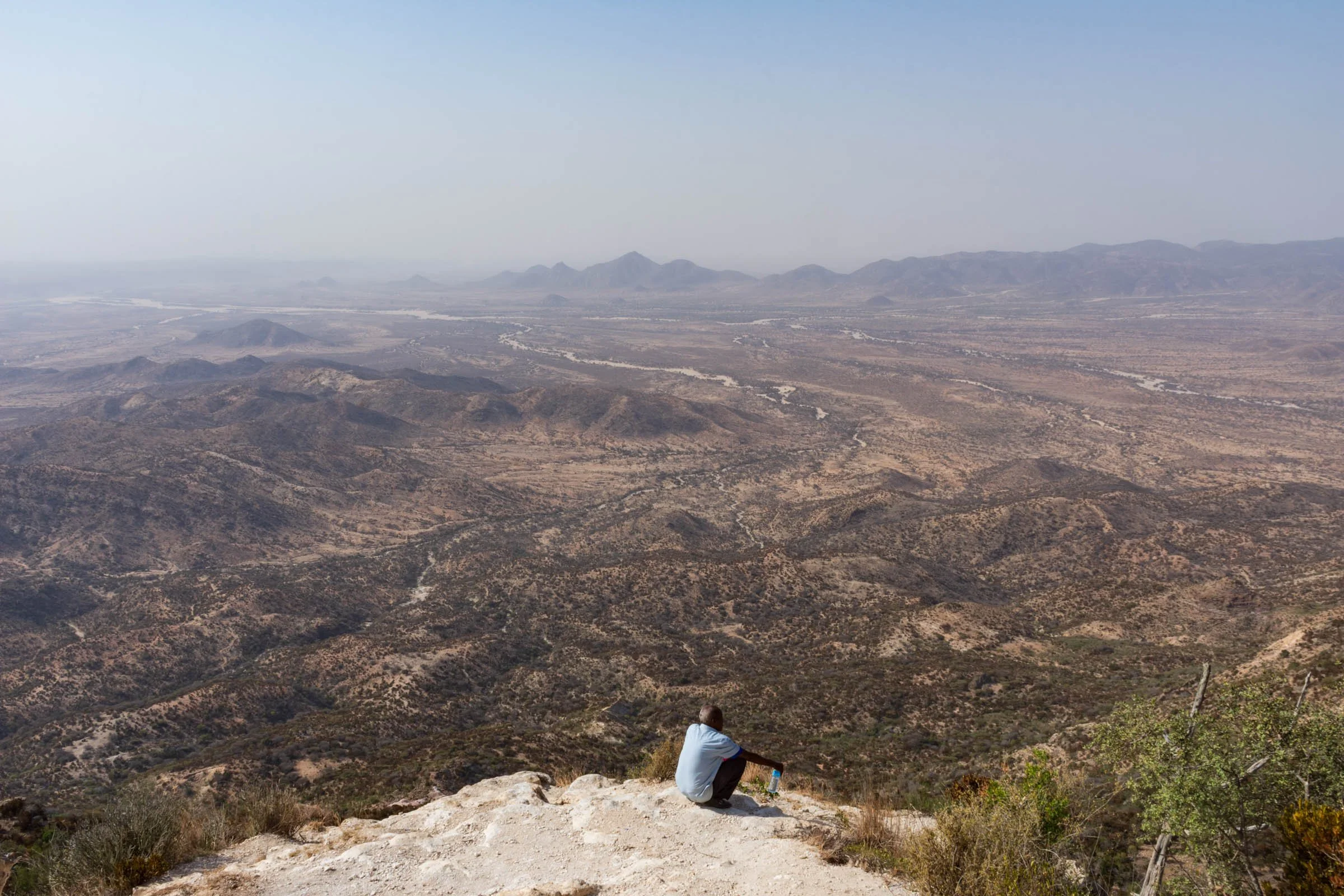 A man sits on the top of a mountain escarpment looking out across the panoramic view over peaks and dry riverbeds