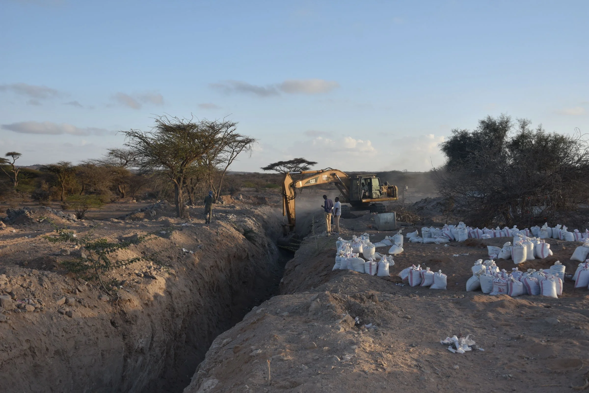 Men watch as an excavator clears a ditch, beside a circle of sandbags