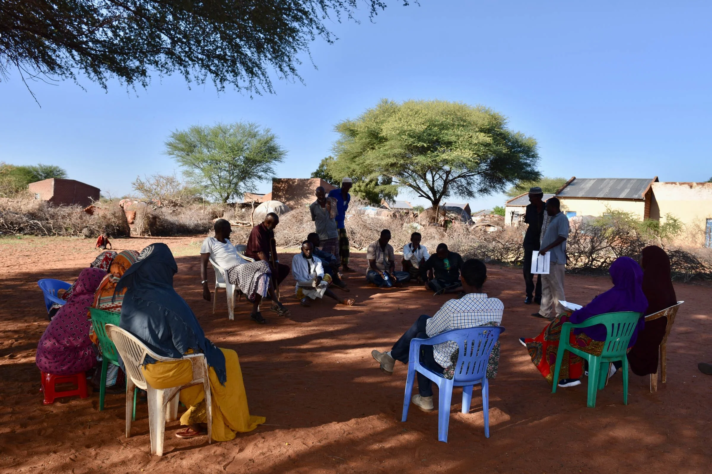 A circle of men and women sit under the shade of trees in a Somali village